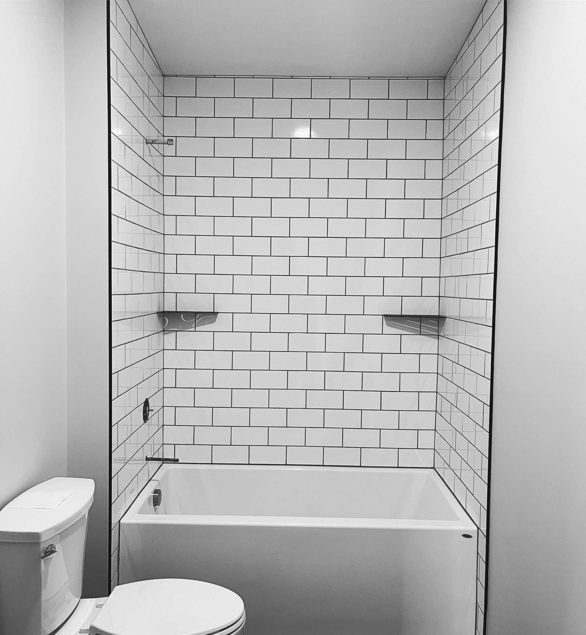 A white bathtub in a shower enclosure with white subway tile walls and two small metal corner shelves.