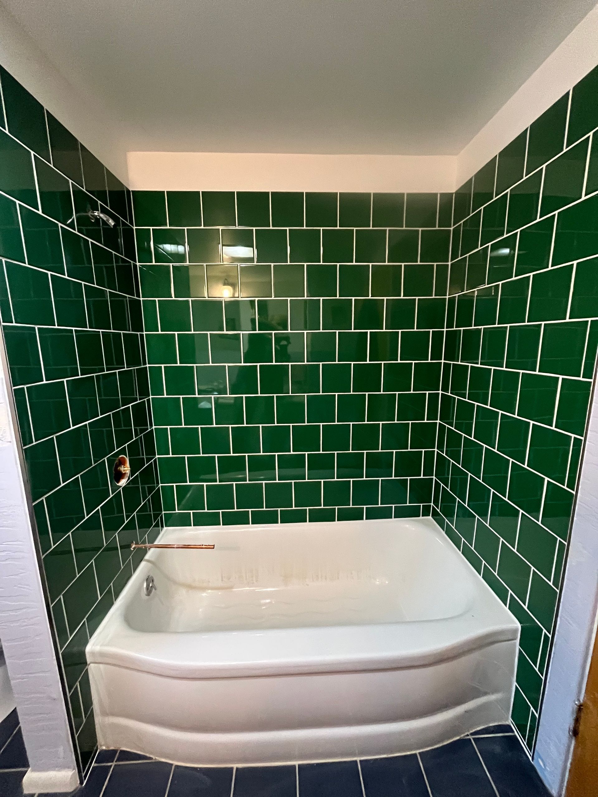 A white bathtub set against walls covered in glossy dark green rectangular tiles with white grout.