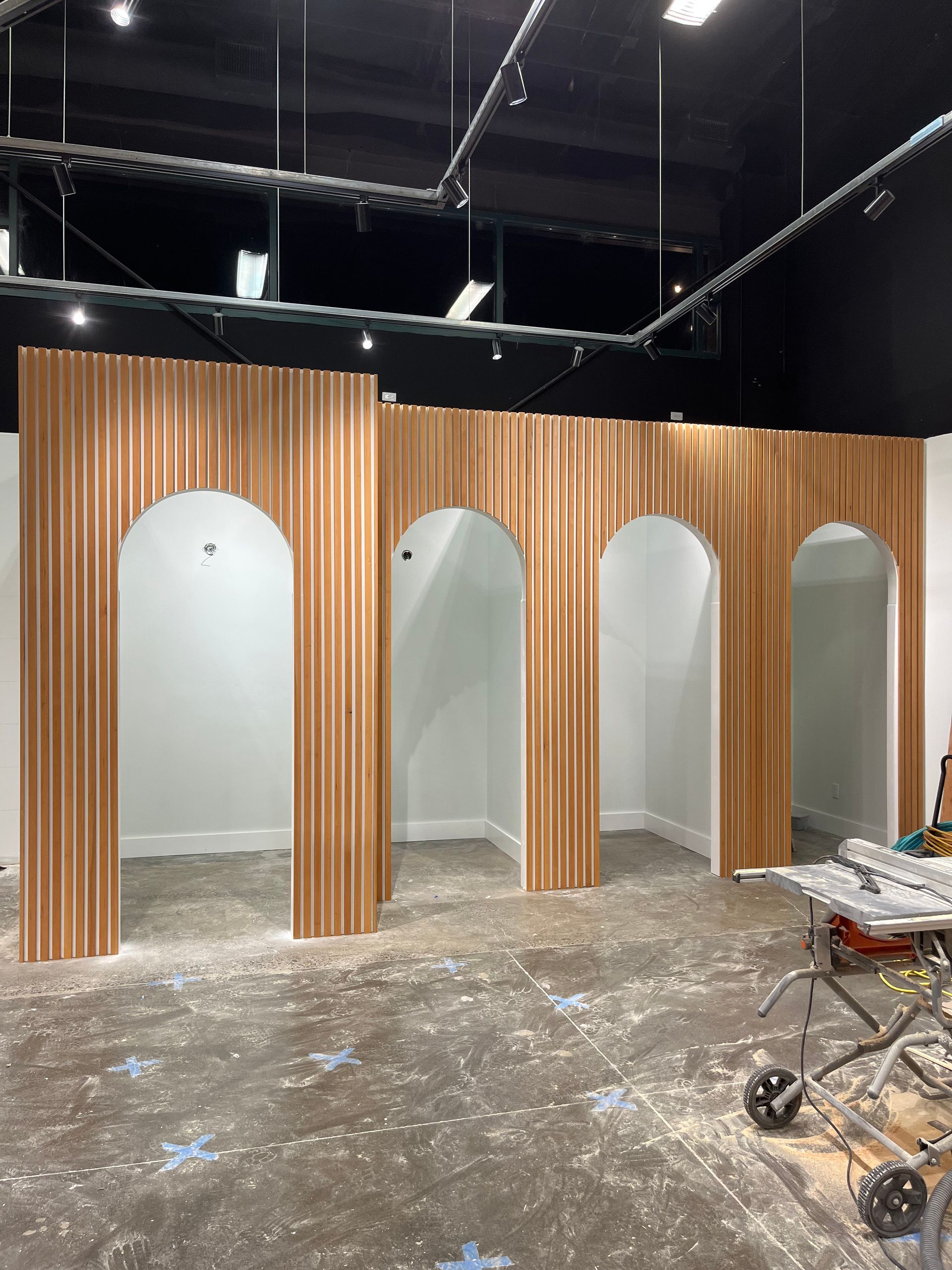 Four arched openings framed by vertical, light-colored wood slats in a room under construction with a dusty floor.