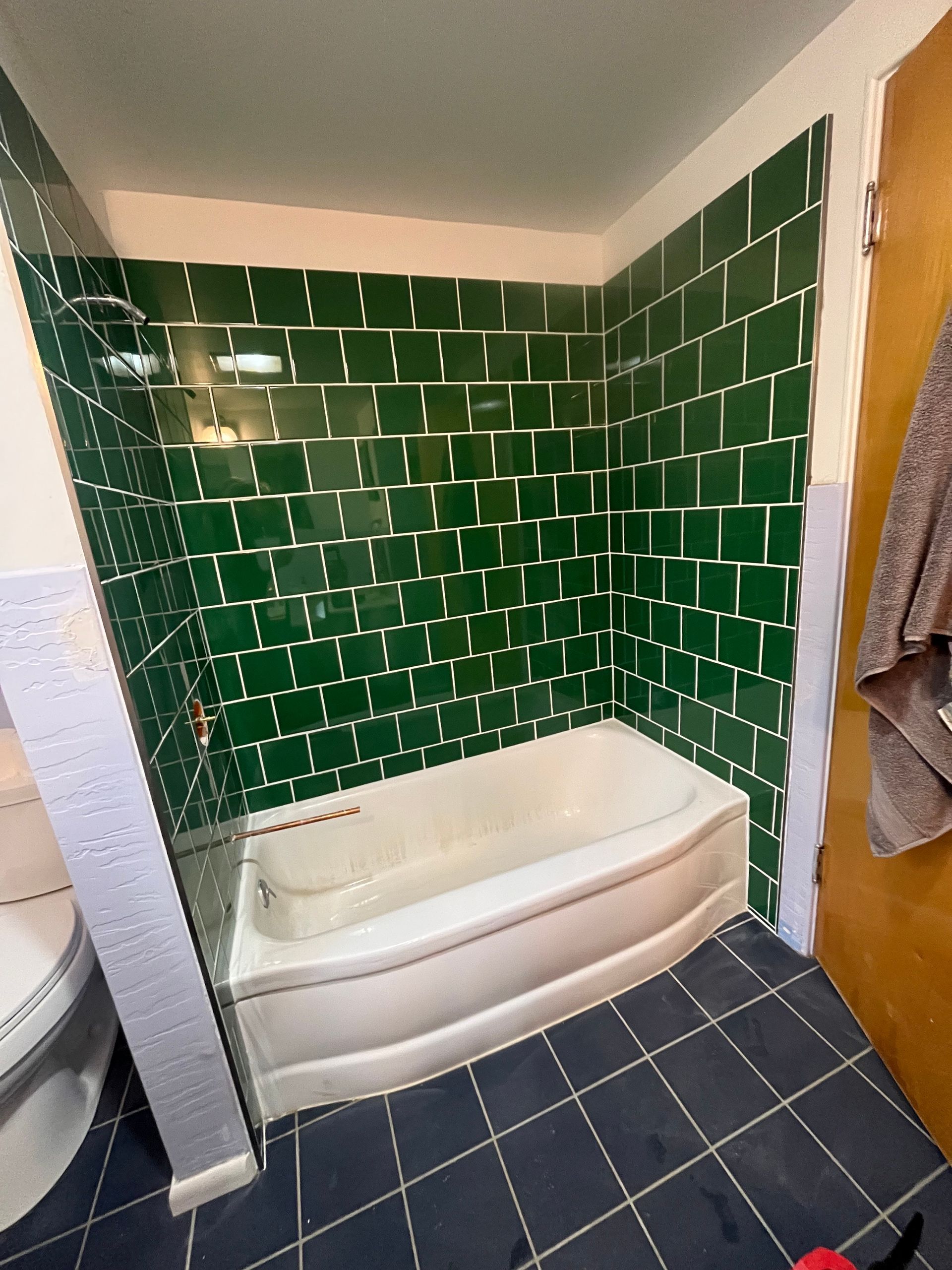 A bathroom with a white curved bathtub surrounded by deep green, rectangular subway-style tiles and dark floor tiles.