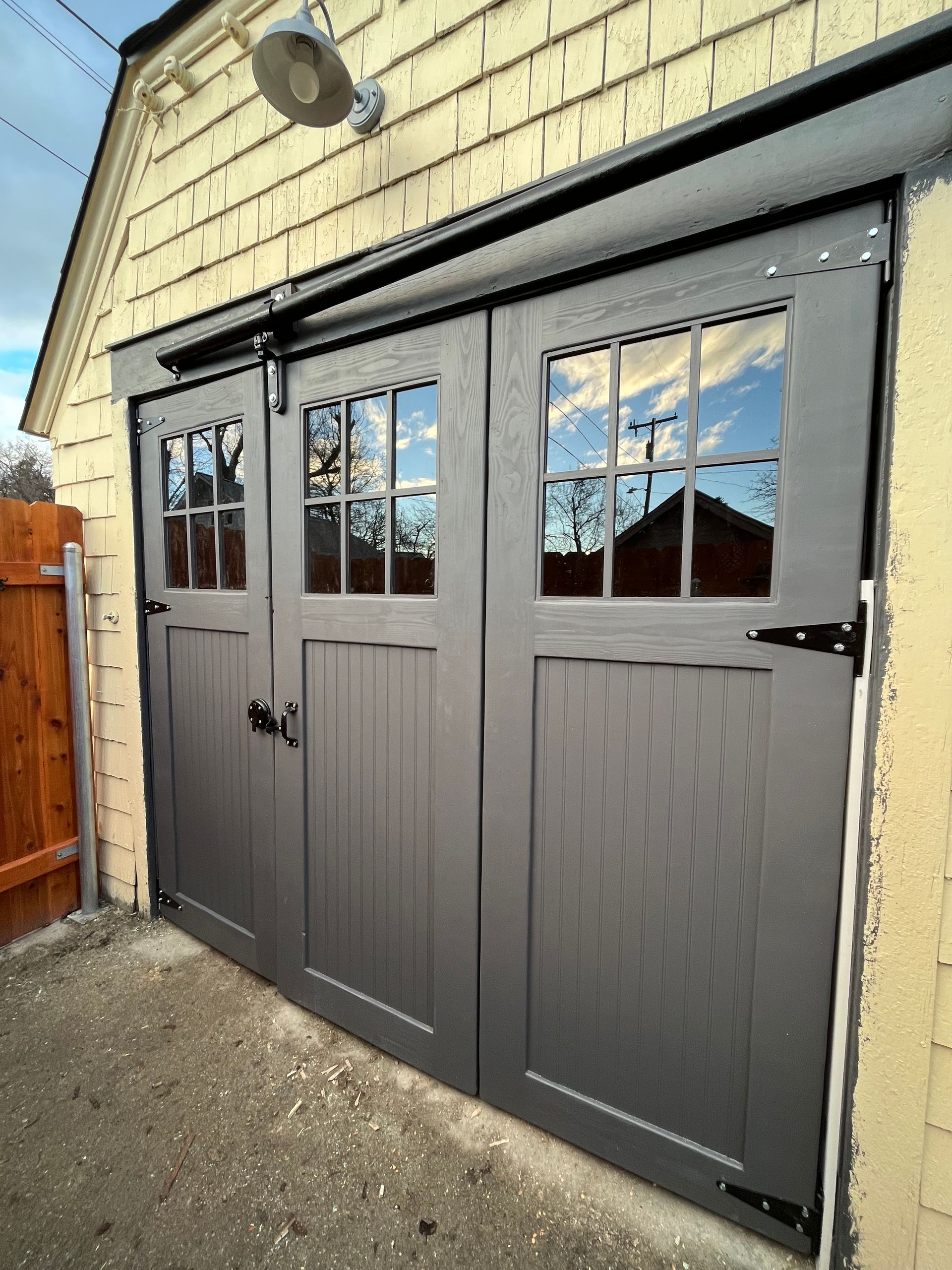 Dark gray, multi-panel carriage-style doors with glass windows on a light yellow shed with a black sliding track and hinges.