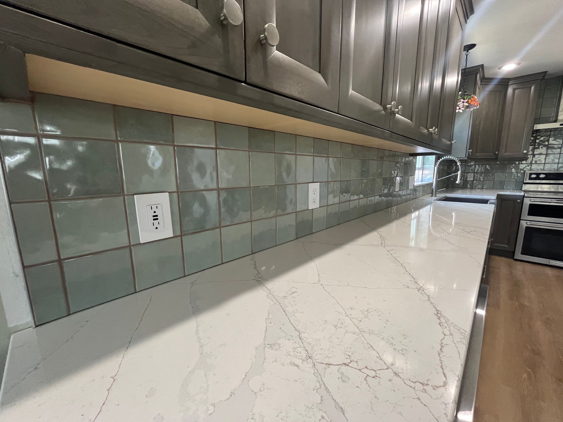 A kitchen backsplash featuring light green square tiles above a white marble countertop with grey cabinets overhead.