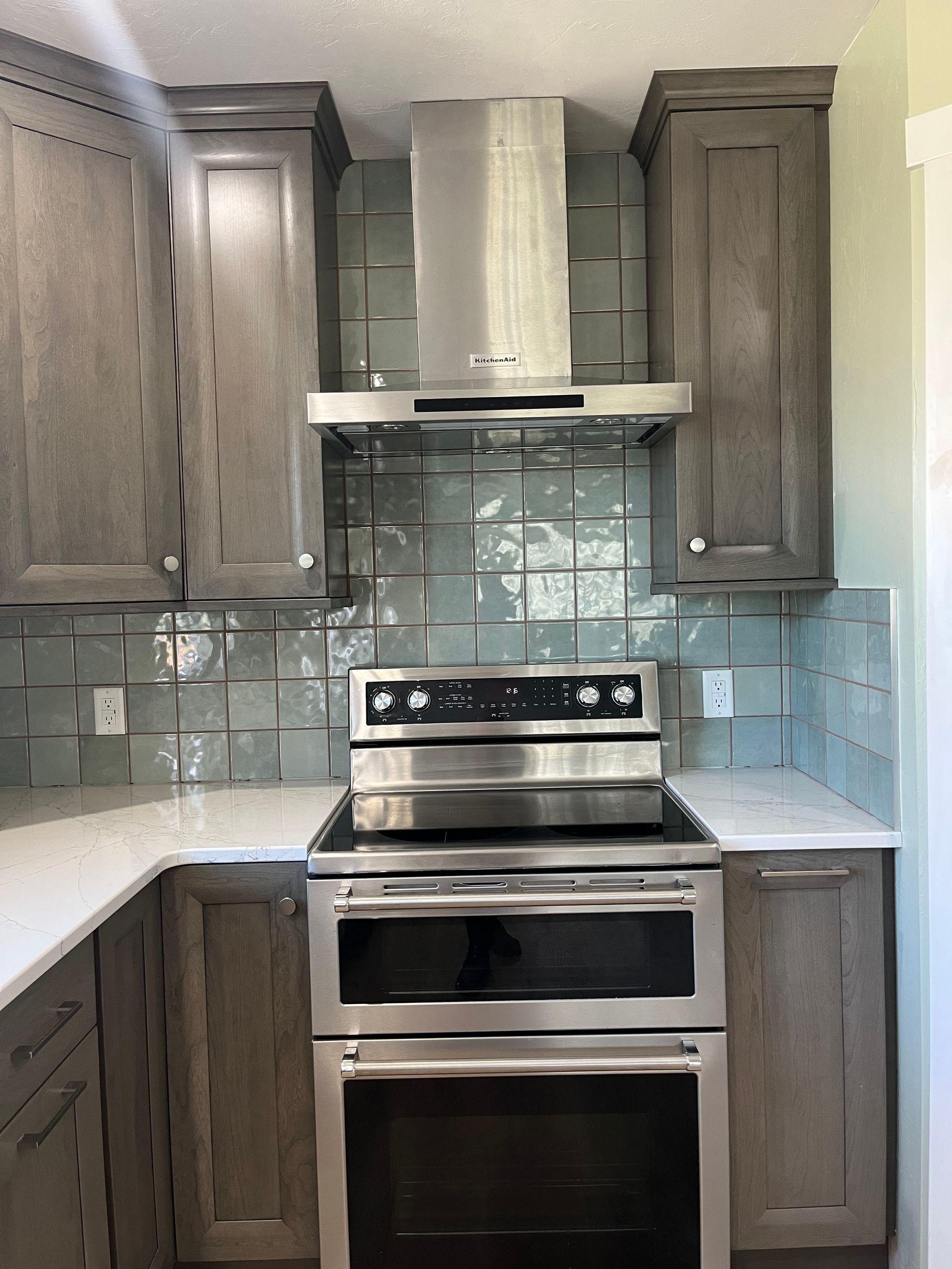 A modern kitchen featuring grey cabinets, a stainless steel stove with double ovens, a range hood, and blue-grey backsplash.
