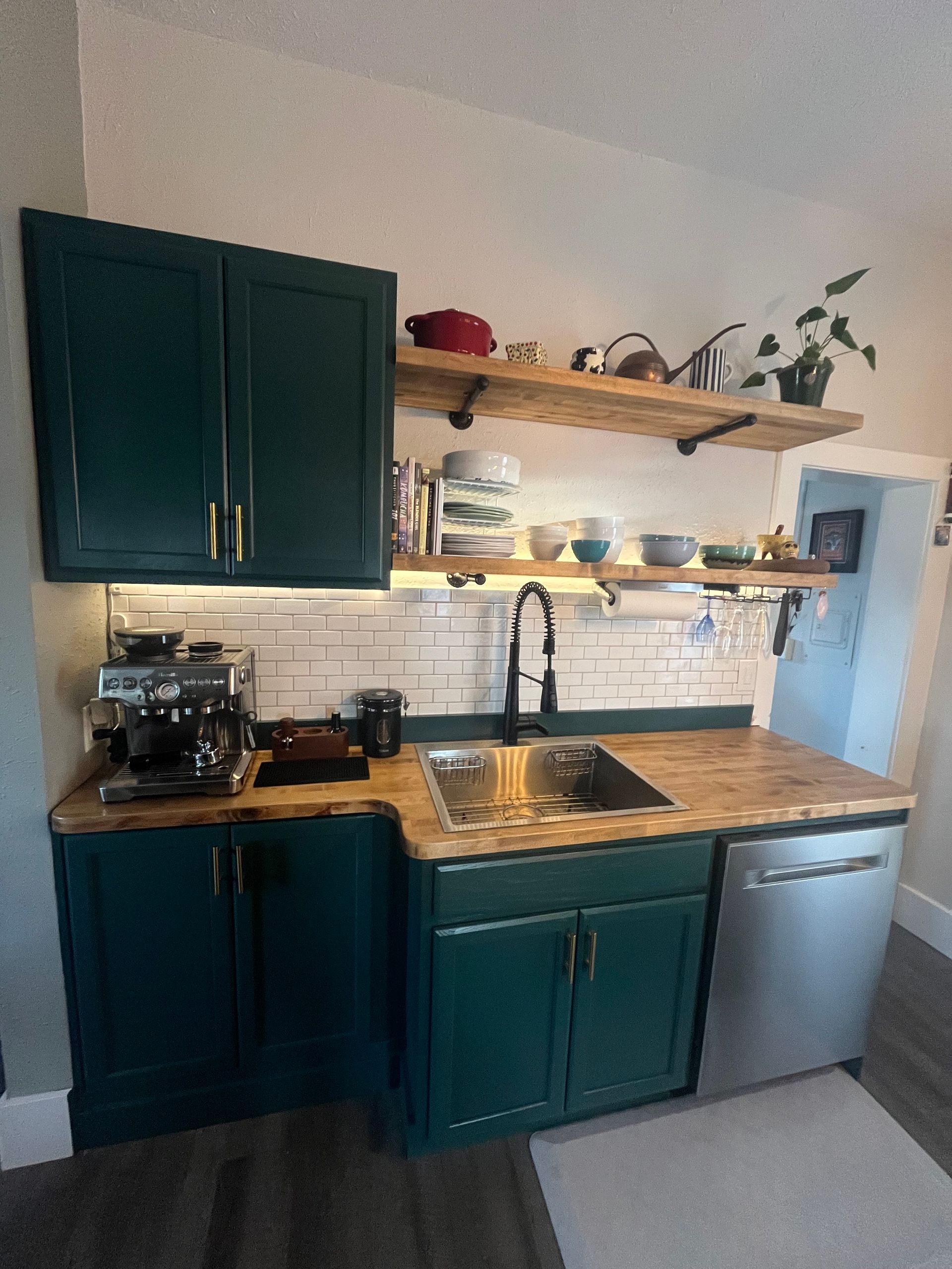 Kitchen counter with dark teal cabinets, a butcher block countertop, two wood floating shelves, and a white subway tile.