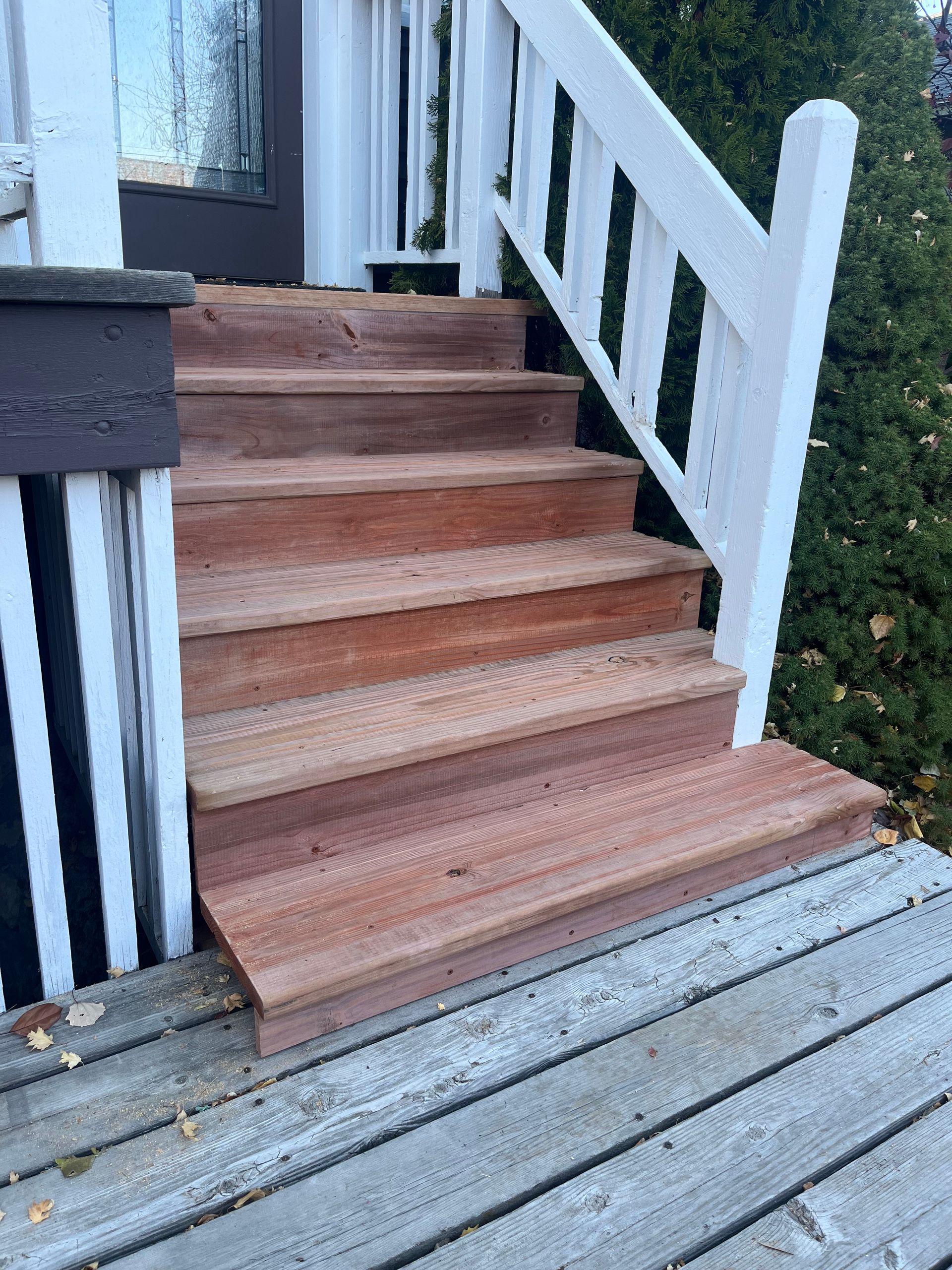 A set of four new natural wood stairs leading to a doorway, flanked by a white railing and an existing weathered deck.