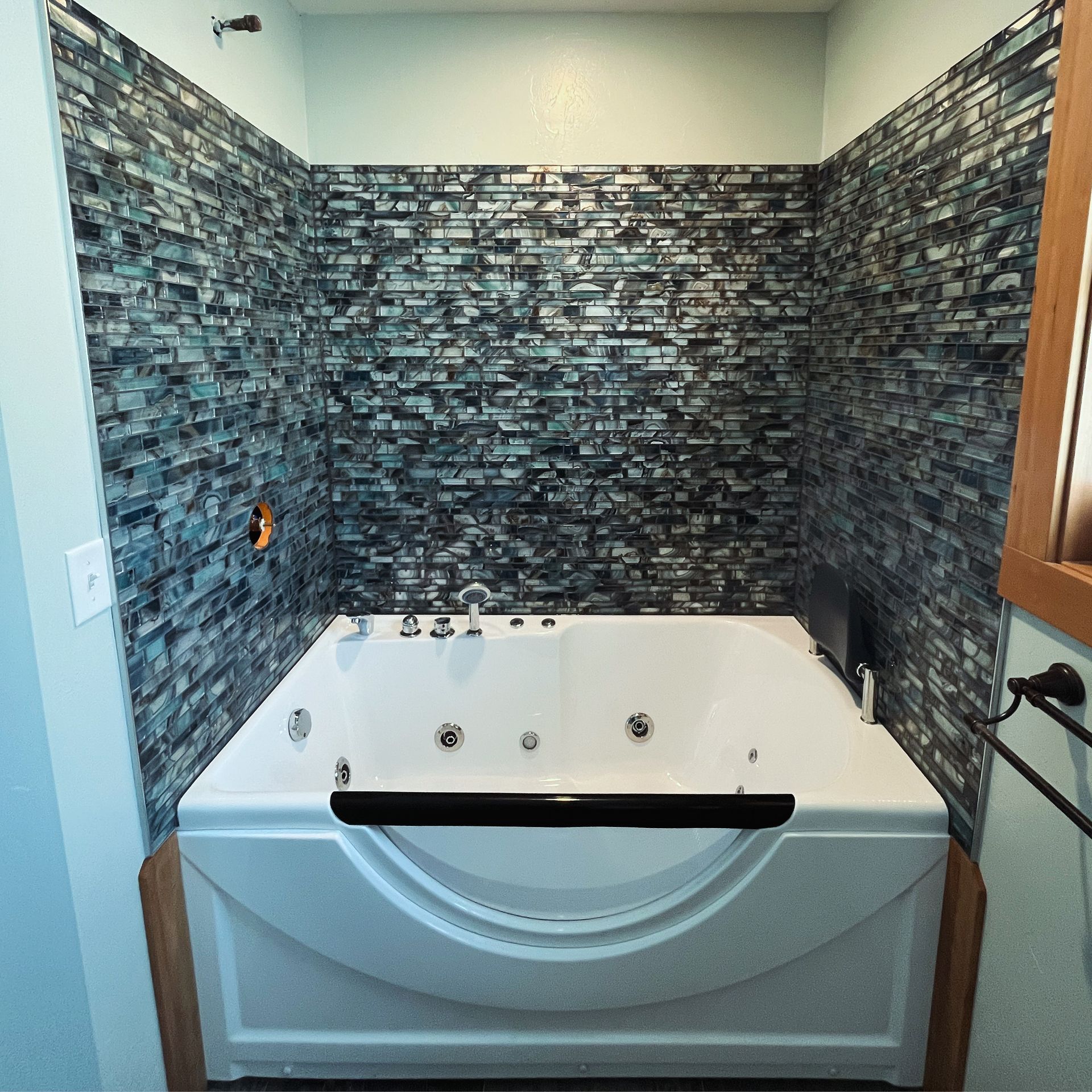 A white jetted bathtub set against a wall featuring dark blue and grey horizontal mosaic tile.