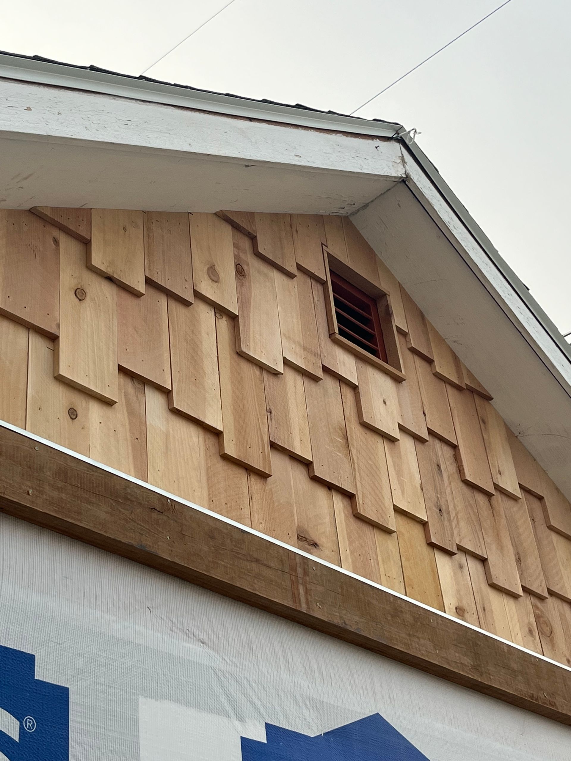 A close-up of a house gable being installed with light wood shingle siding and a small, square brown attic vent.