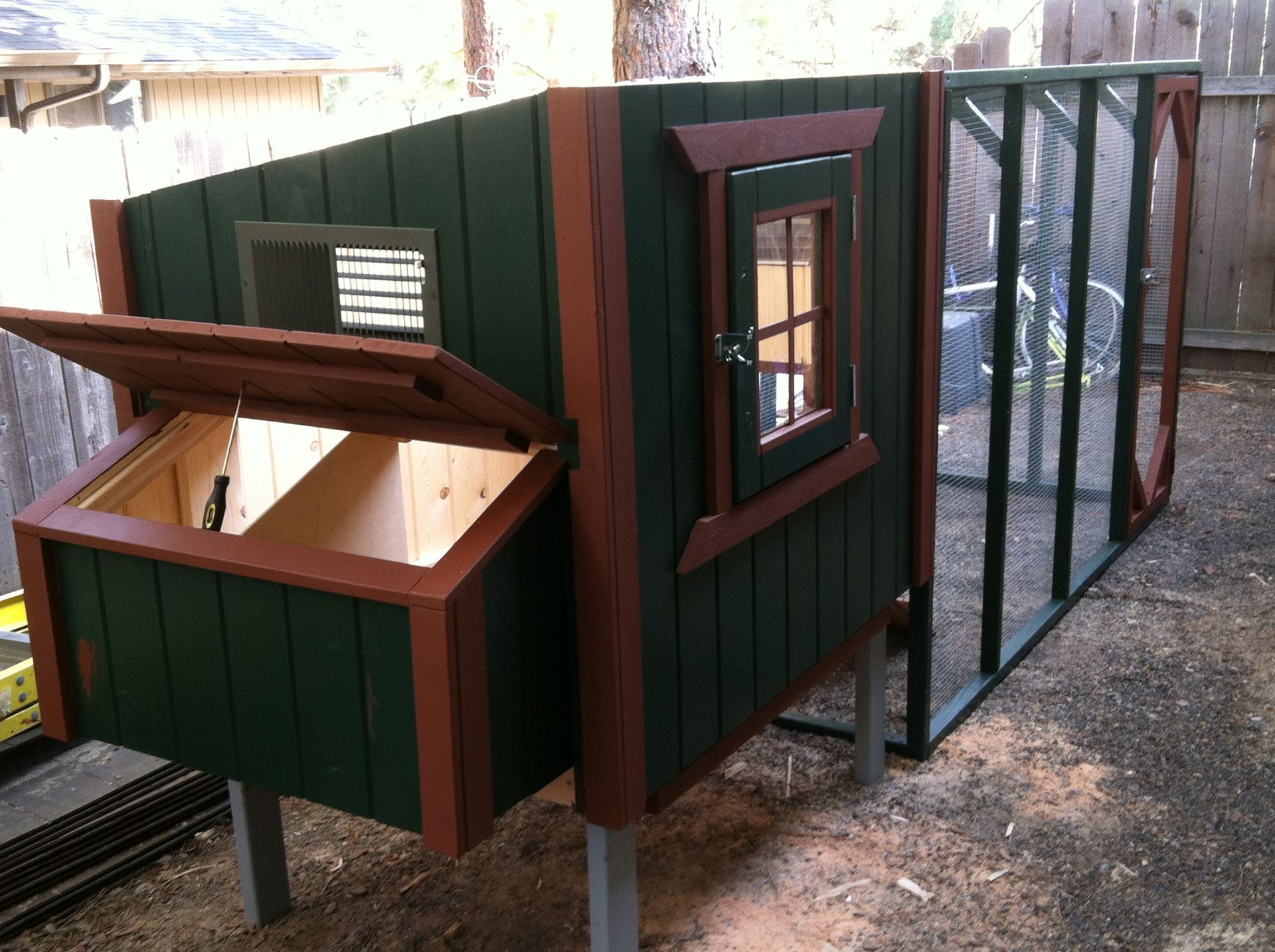 A green and brown wooden chicken coop with an open nesting box and attached wire run in a backyard.