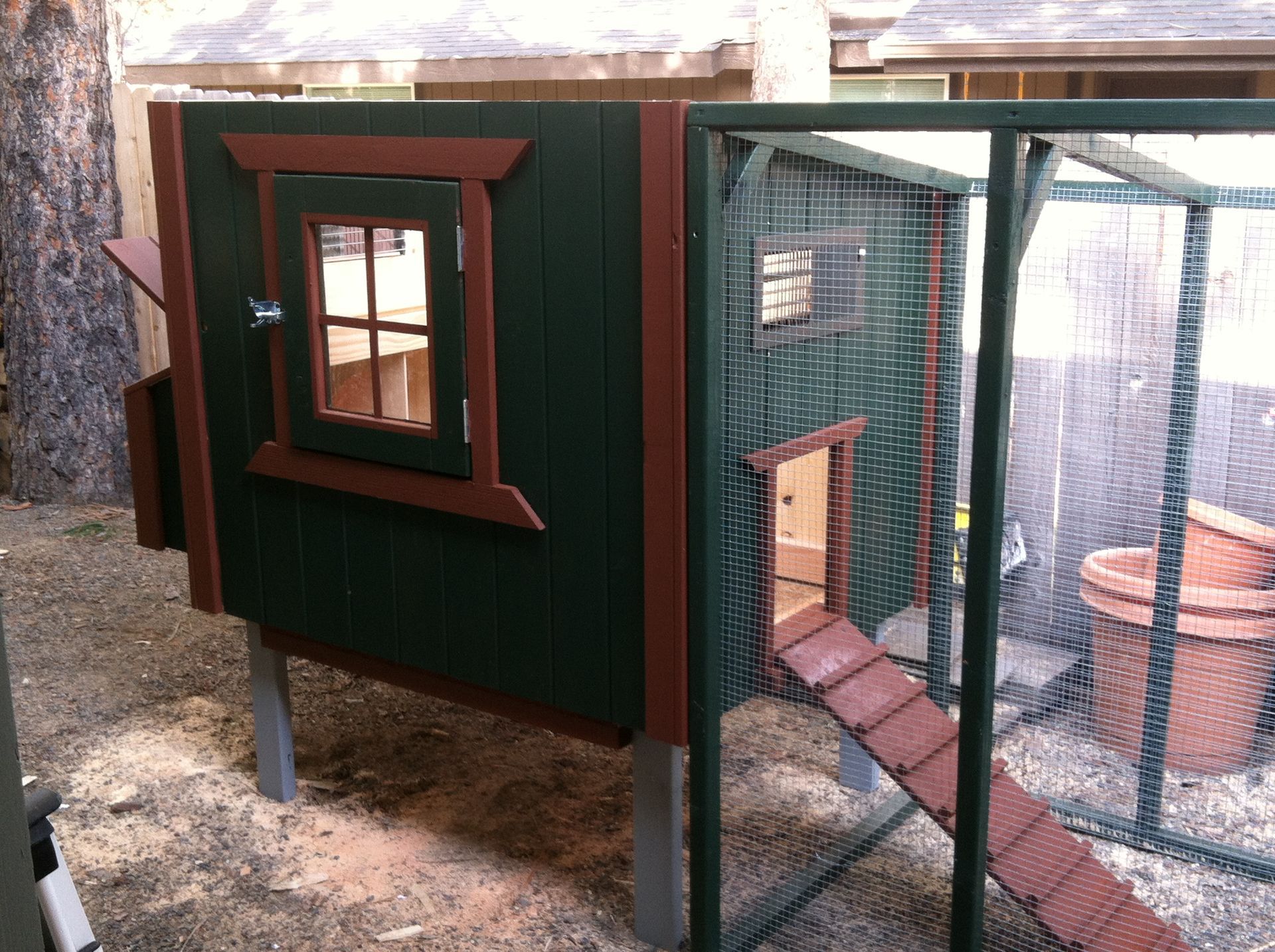 A dark green and brown wooden chicken coop with a window and a small ramp leading into an attached wire enclosure.
