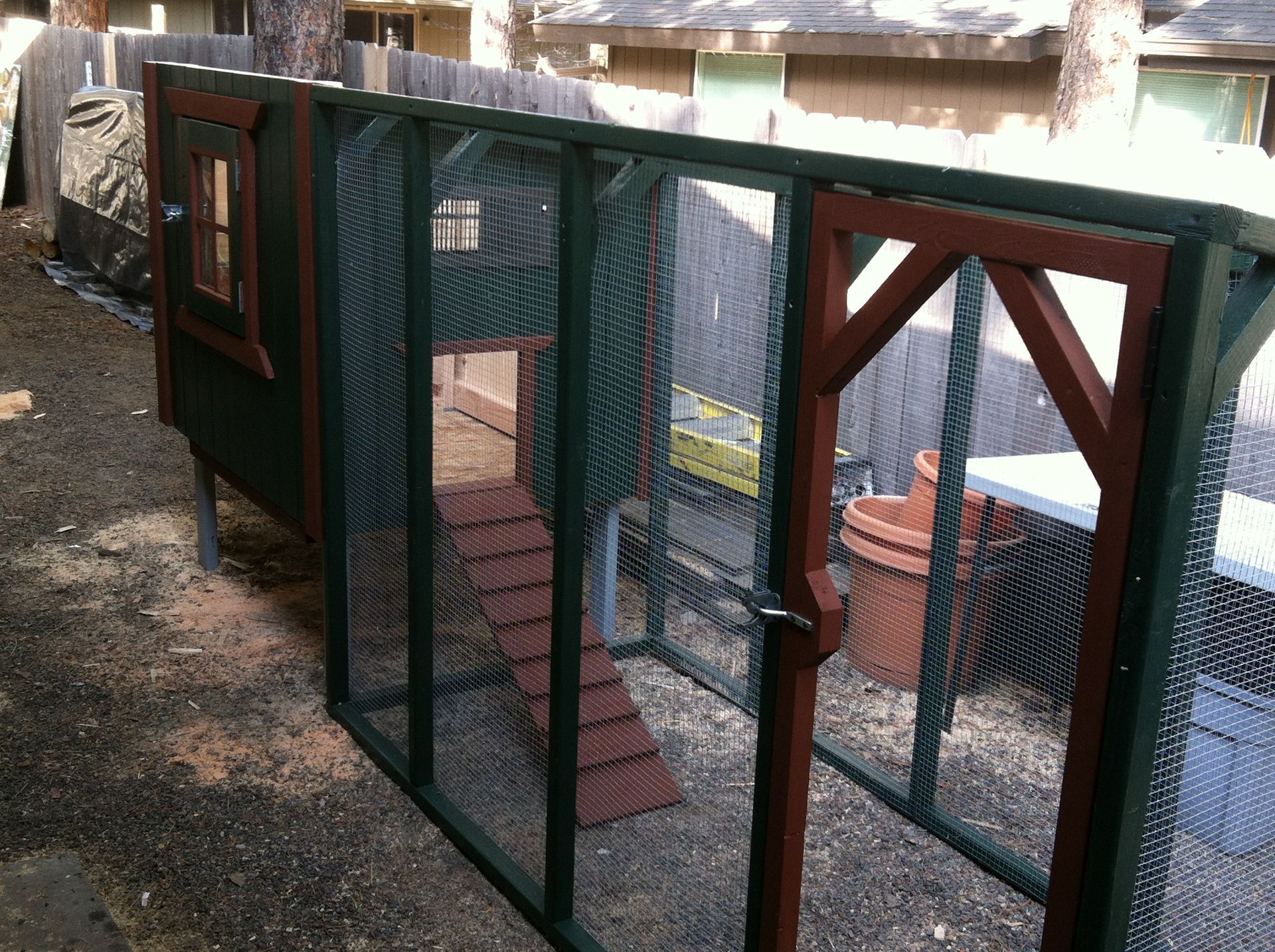 A dark green and brown wooden chicken coop with a wire mesh run and a ramp, situated in a backyard.