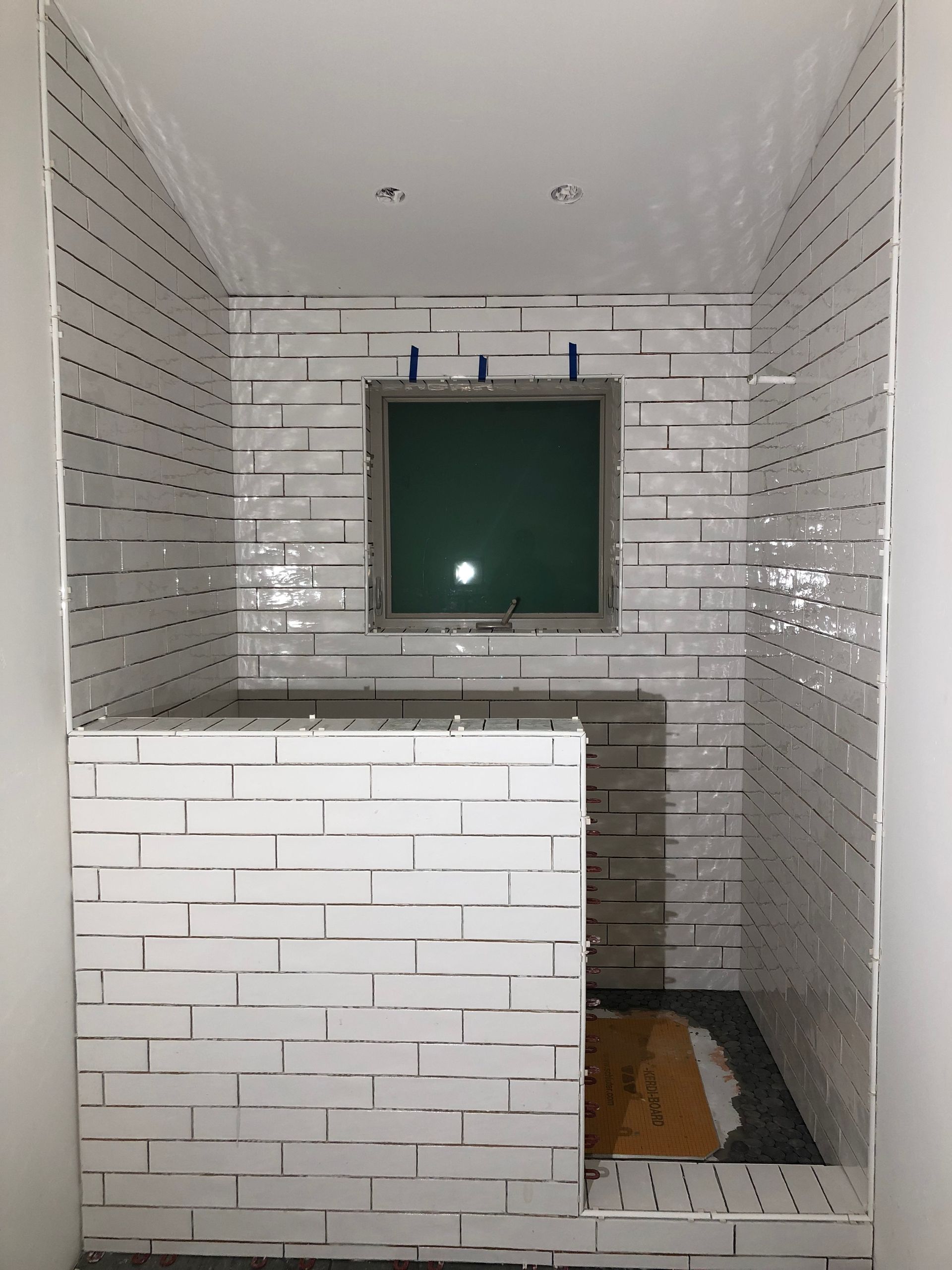 A shower under construction with white subway tile on the walls and a low, tiled partition wall in a bathroom.