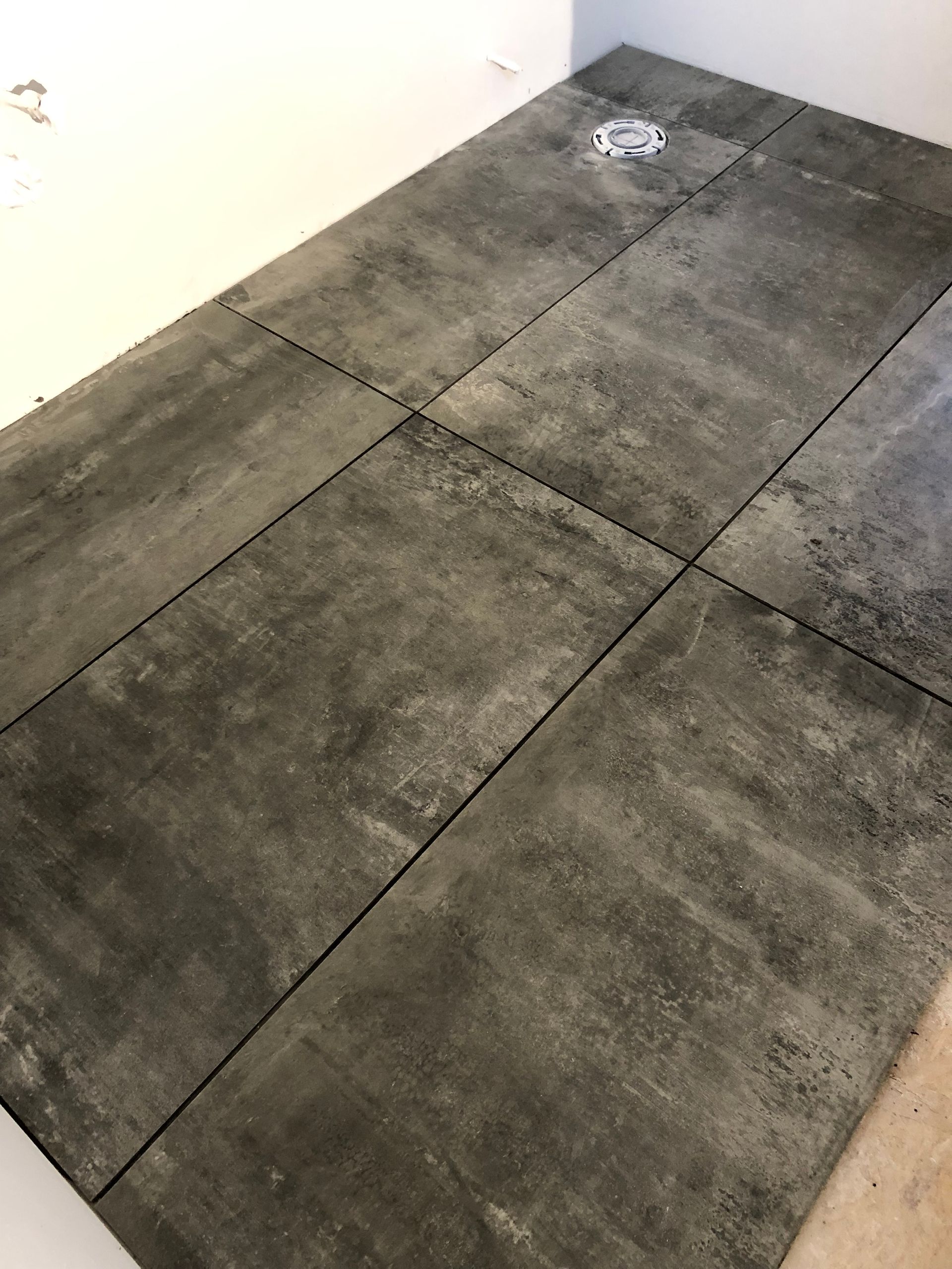 Large, dark gray, rectangular floor tiles installed in a bathroom, with a circular drain visible in the upper corner.