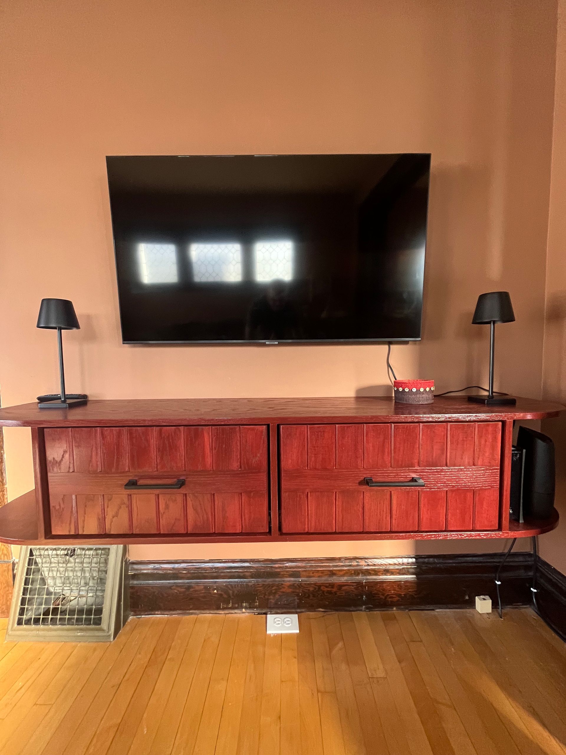 A flat-screen TV mounted above a custom-built, reddish-brown wooden floating media console with two drawers and two lamps.