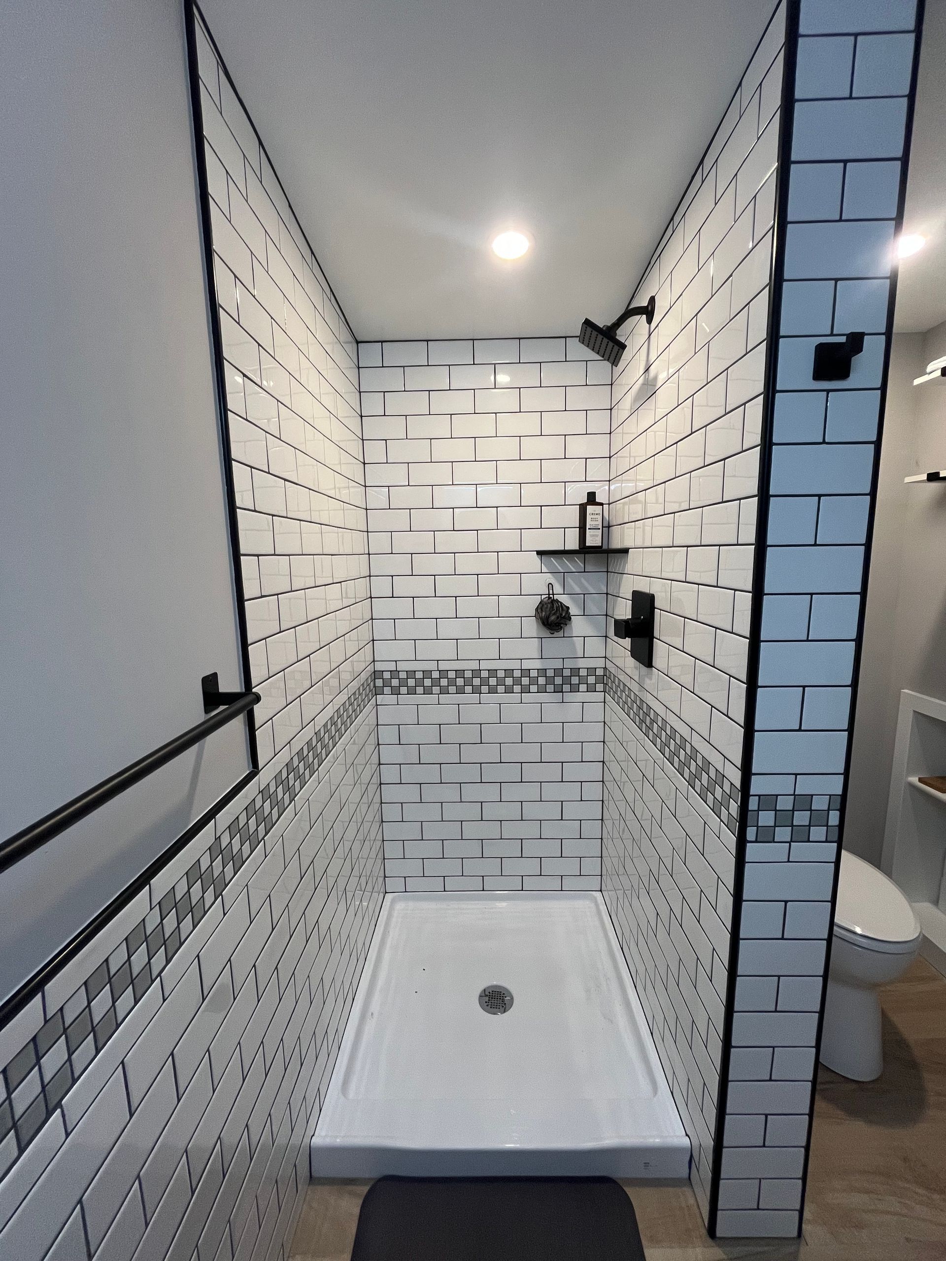 A modern, narrow walk-in shower with white subway tiles, black grout, and a black shower head, next to a white toilet.