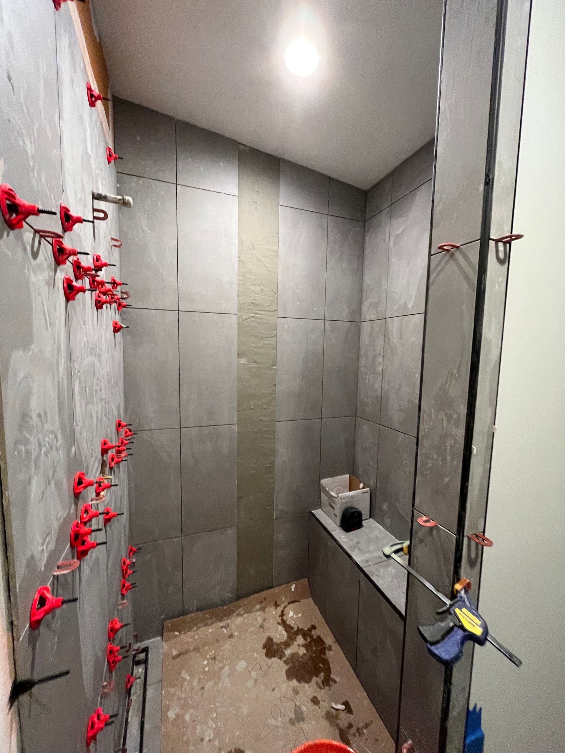 A shower stall under construction with gray wall tiles, red tile-leveling clips, and a built-in bench.