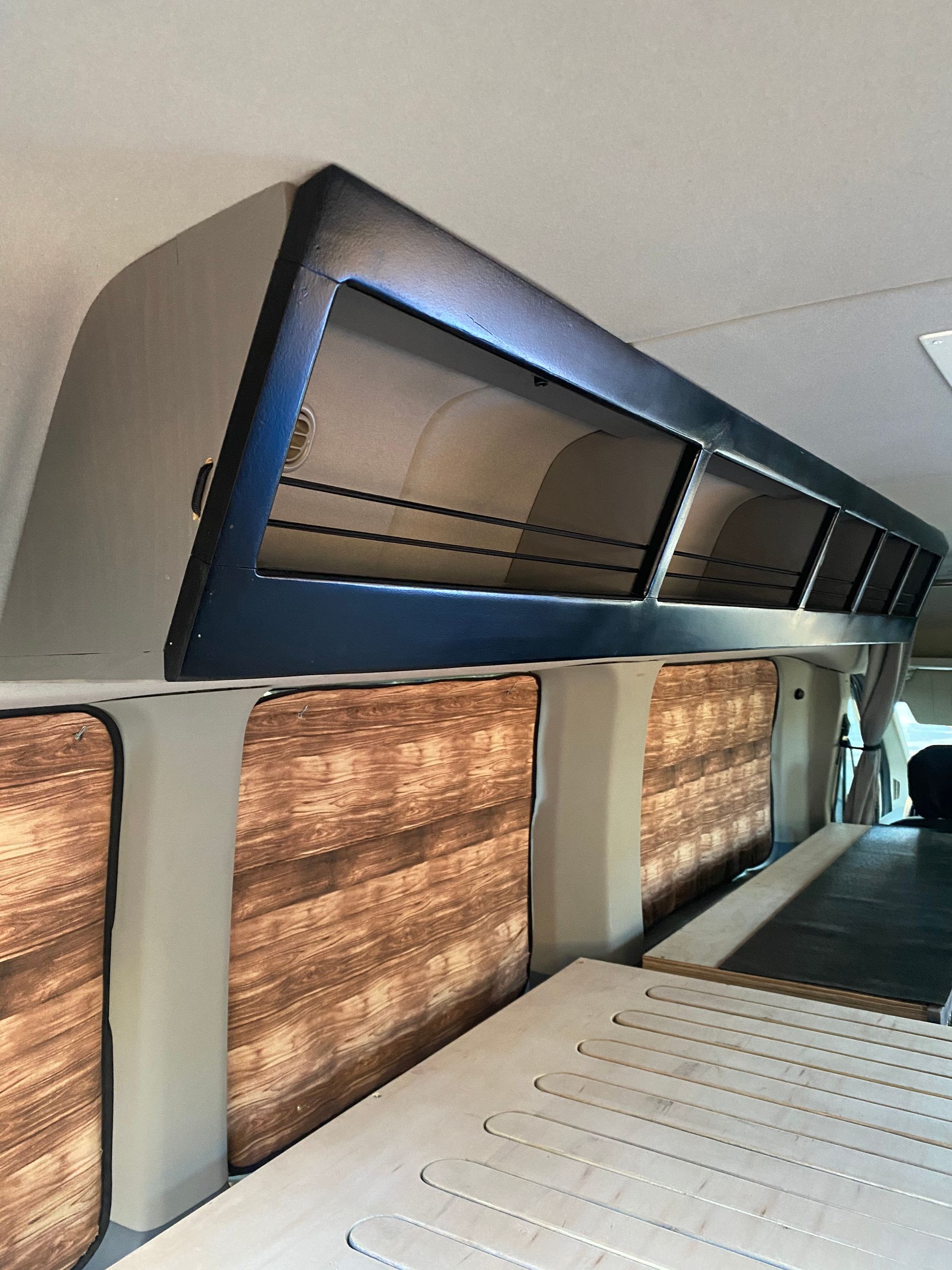 Inside a camper van, a long, black storage cabinet with shelves is mounted above windows covered by wood-patterned blinds.