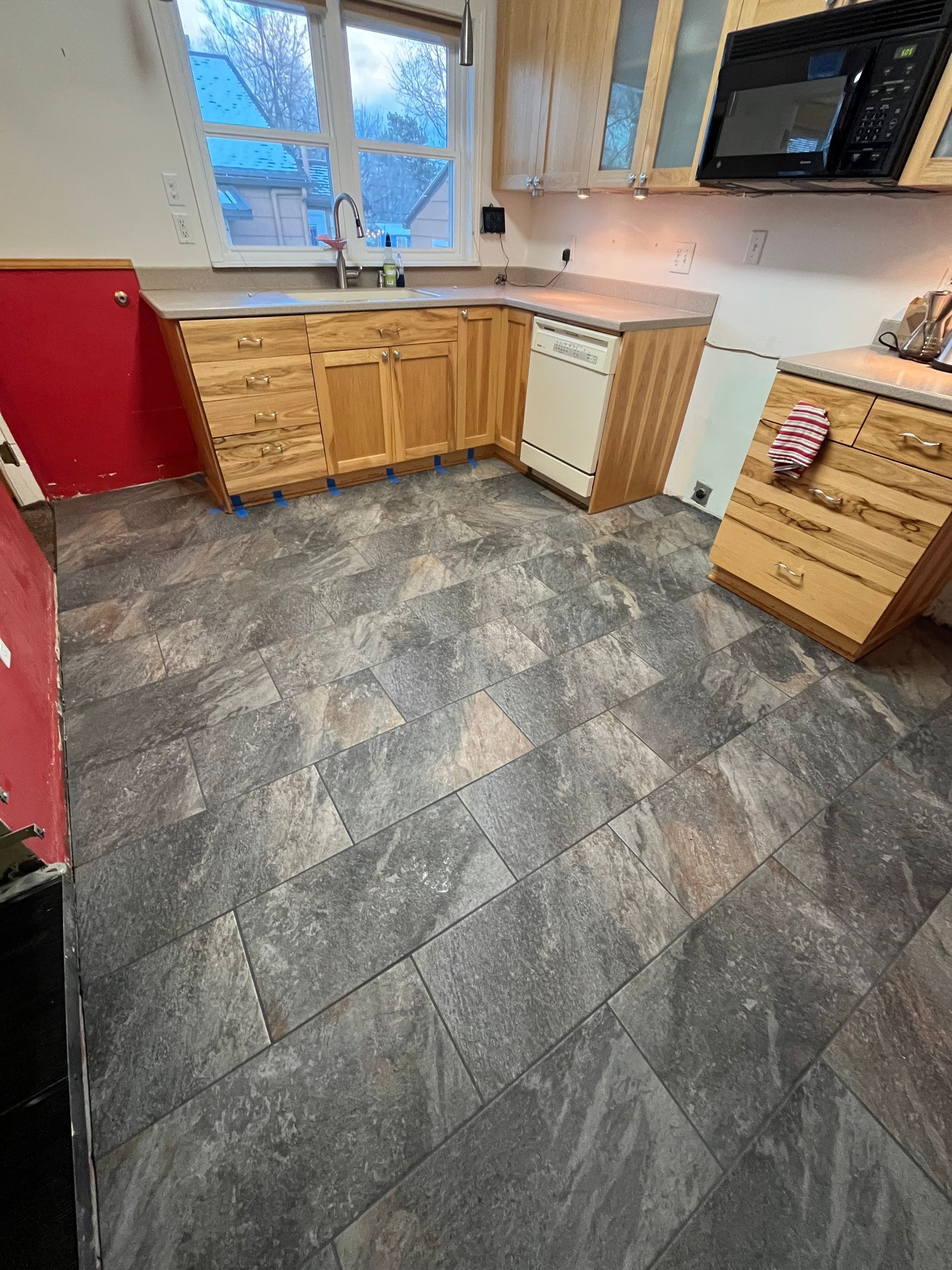 A kitchen with natural wood cabinets, a black microwave, and dark, textured, rectangular floor tiles in a grid pattern.