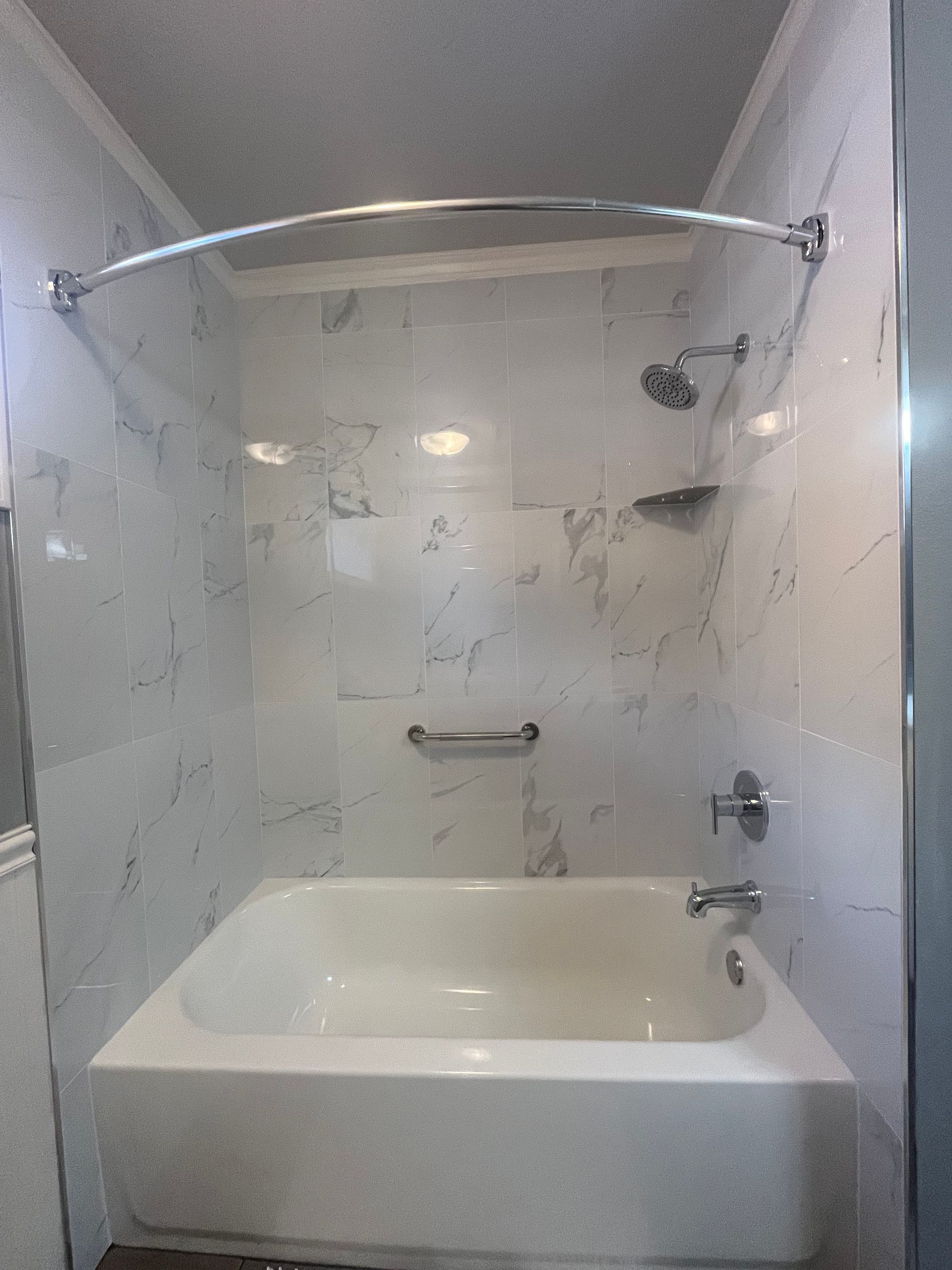 A white bathtub with white marble-patterned tiled walls, a curved shower rod, a grab bar, and chrome plumbing fixtures.