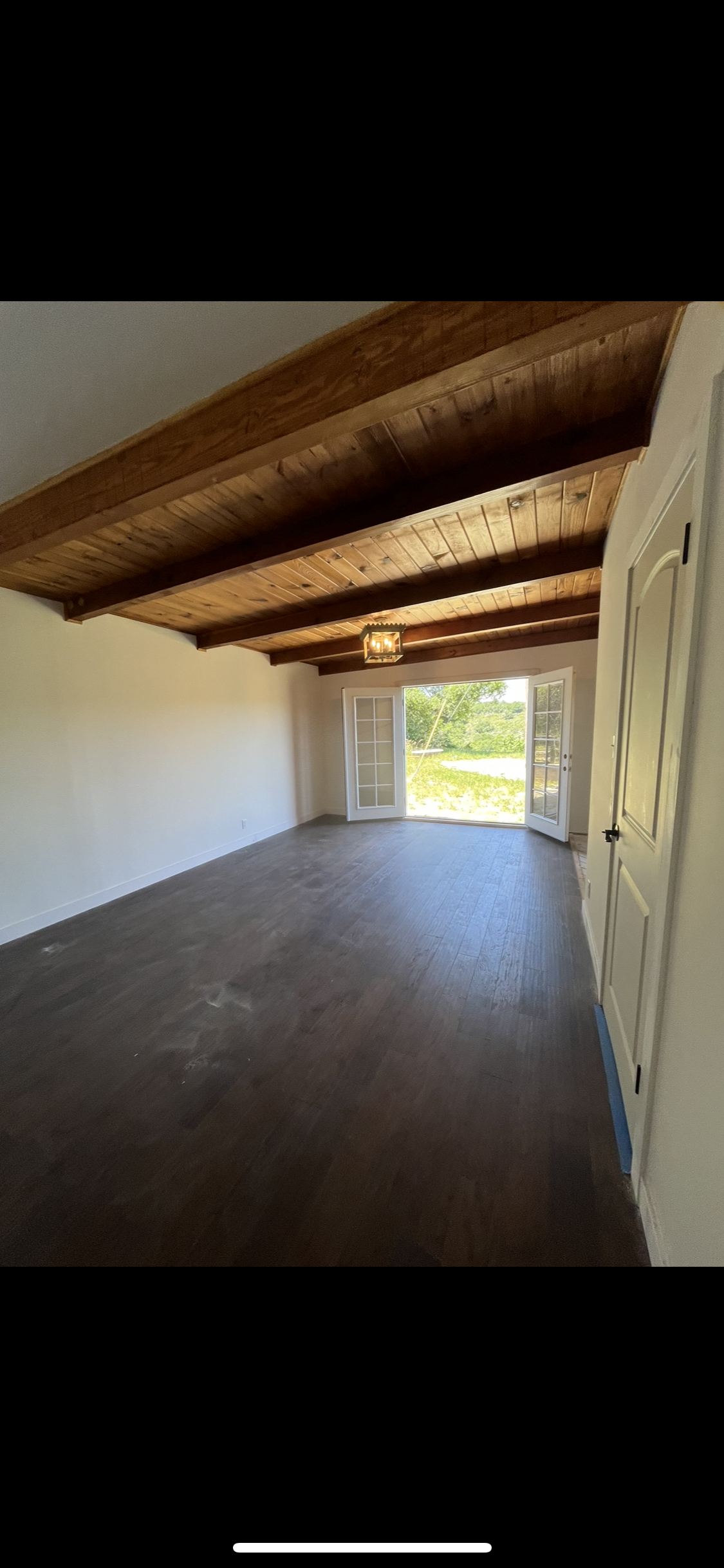 A room with dark wood flooring, white walls, exposed wooden ceiling beams, and French doors leading to an outdoor area.