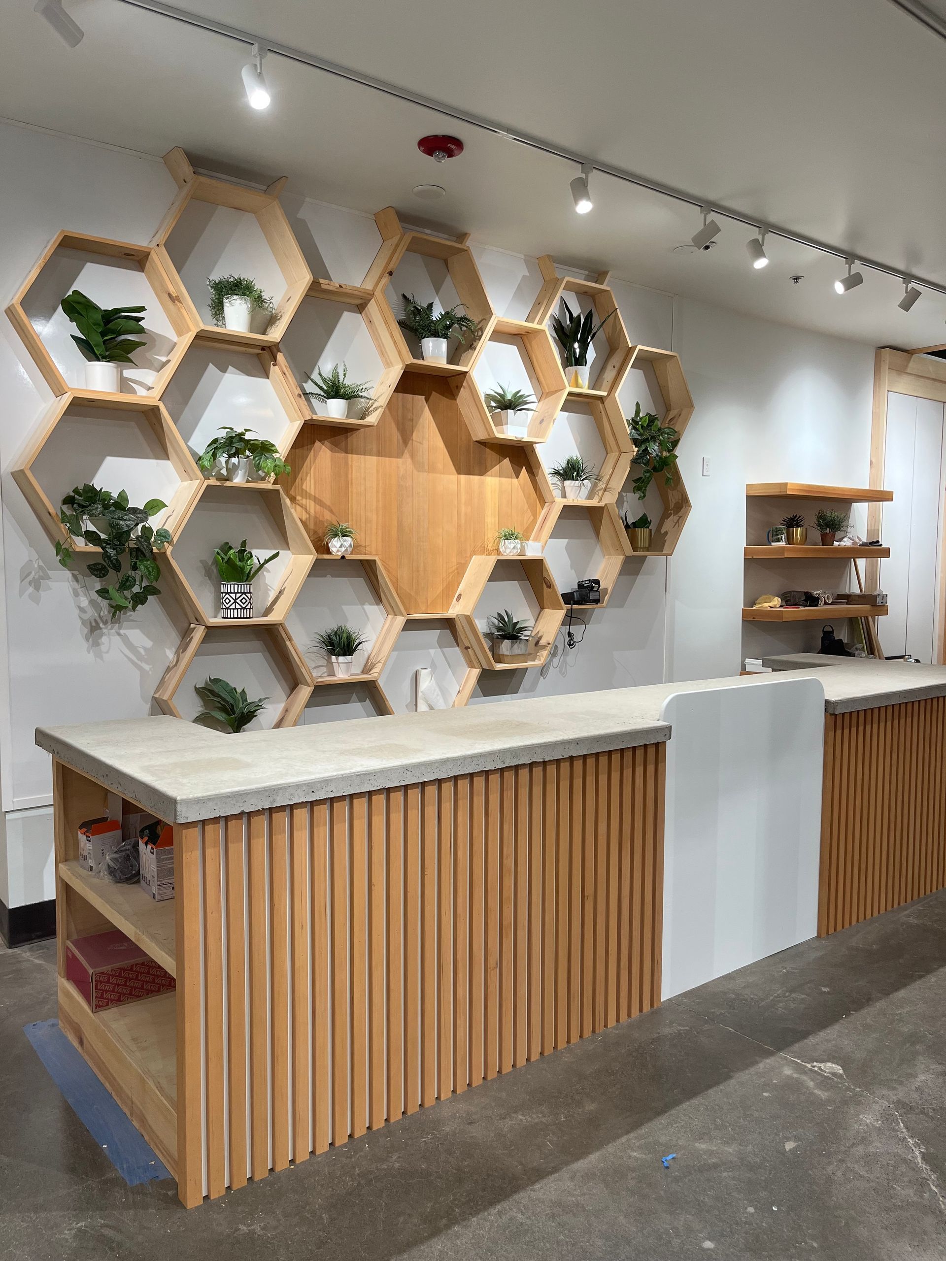 A service counter with a slatted wood base and concrete top, backed by a large wall display of hexagonal wood shelves.