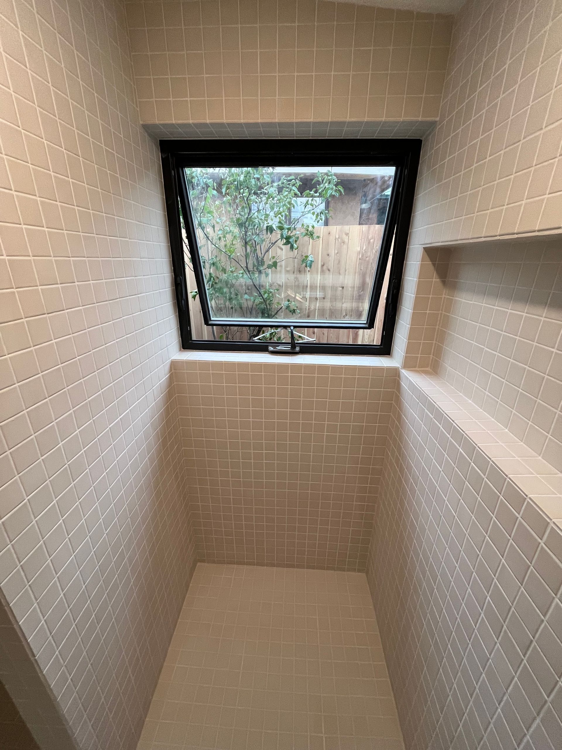 A shower stall with light beige, square-tiled walls, a built-in recessed shelf, and an open, hopper-style window.