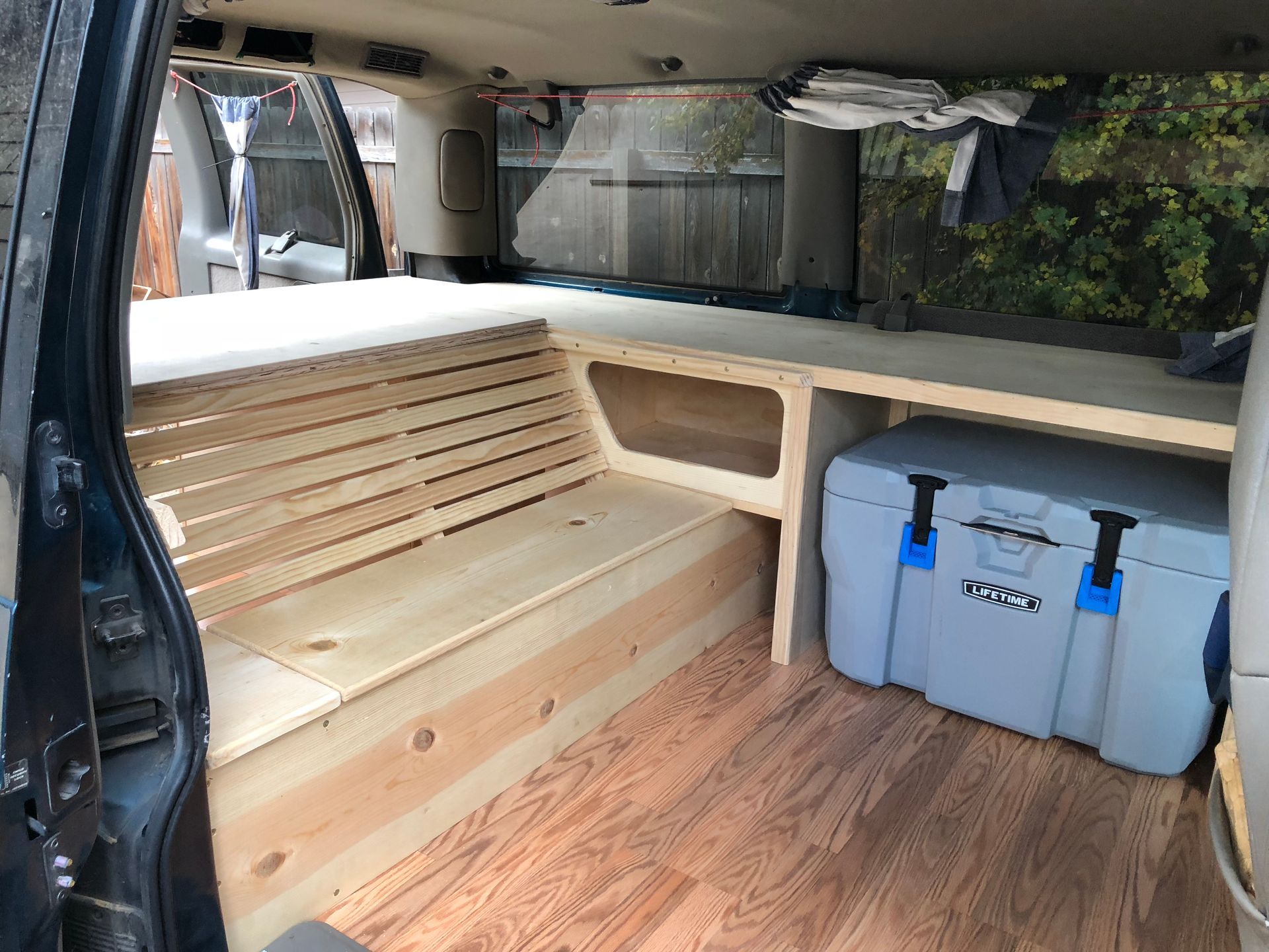 Custom wooden van conversion interior with a slatted bench, shelving, and a grey cooler on a wood-look floor.