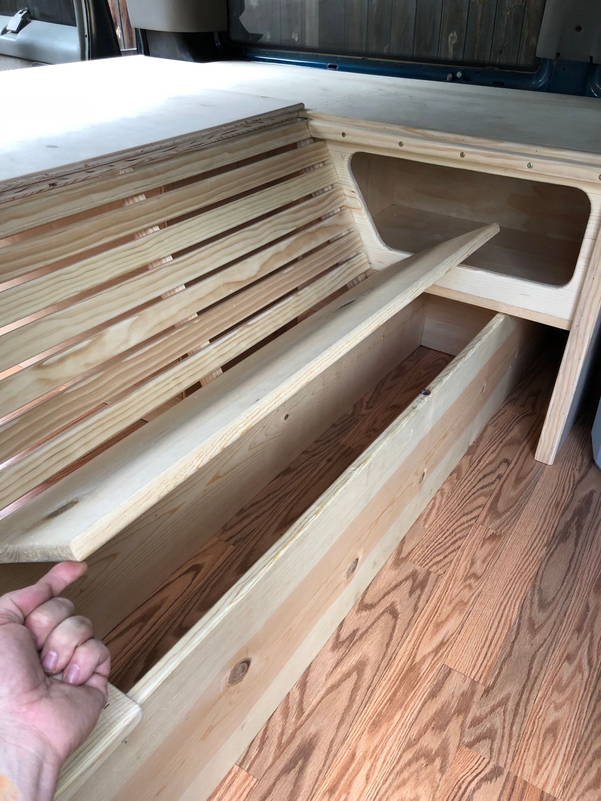 A hand lifts a wooden slat from a custom bed frame being built inside the back of a van with a wood-look floor.