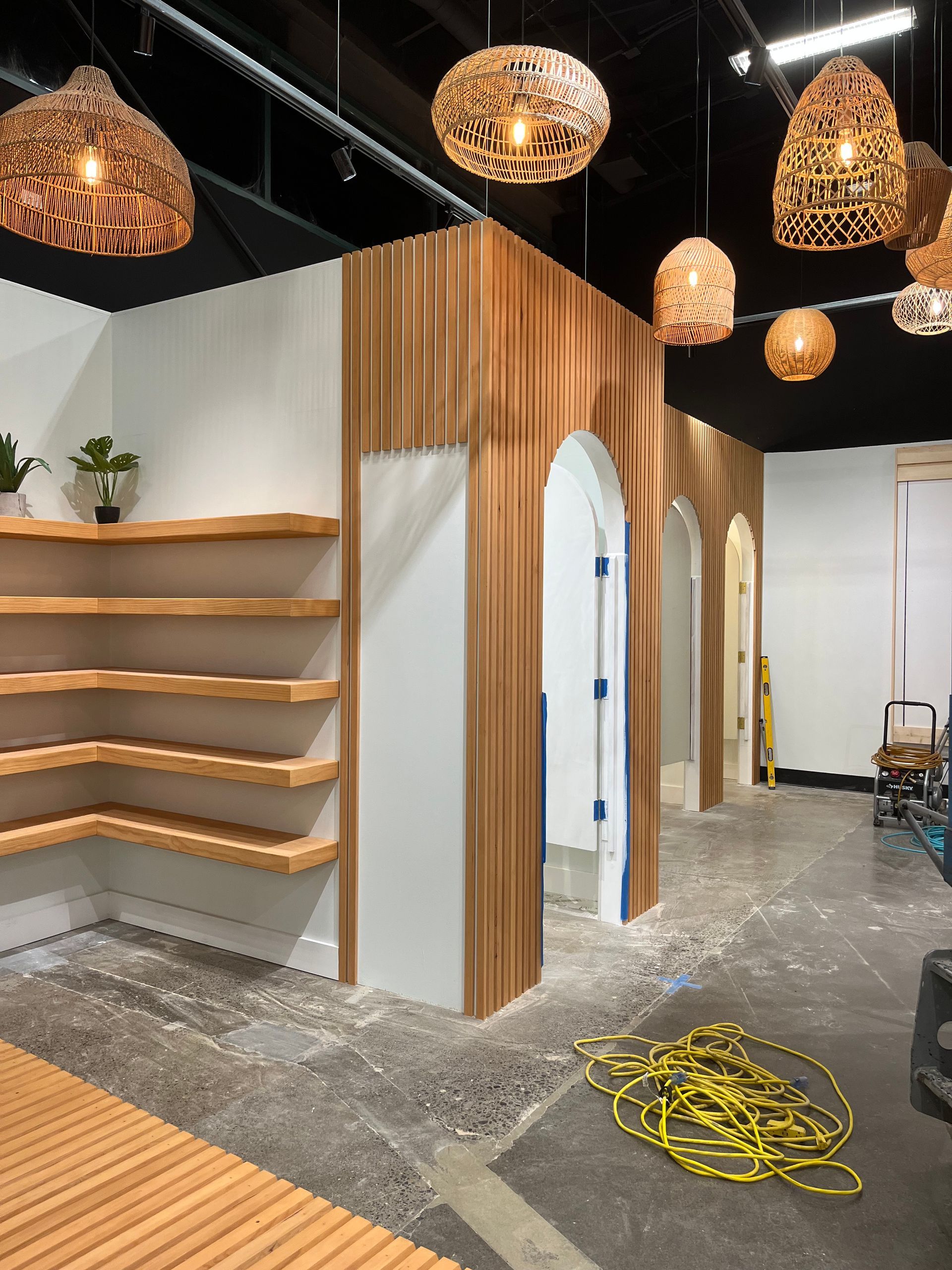 A partially finished retail space with wooden slat walls, arched doorways, corner floating shelves, and hanging lights.