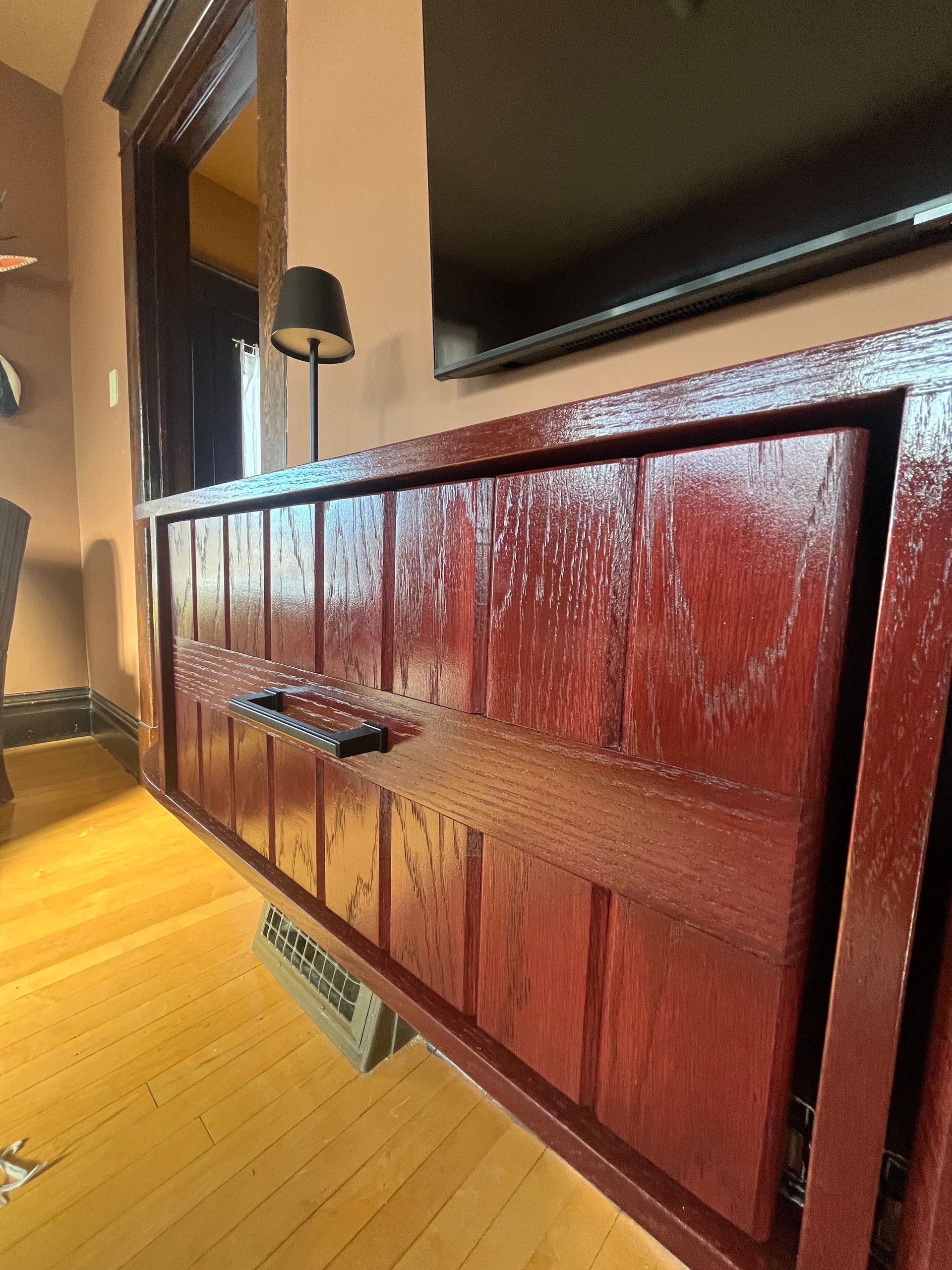 A shiny, deep-red wooden cabinet with vertical panels and a long black handle sits in a room with hardwood floors.