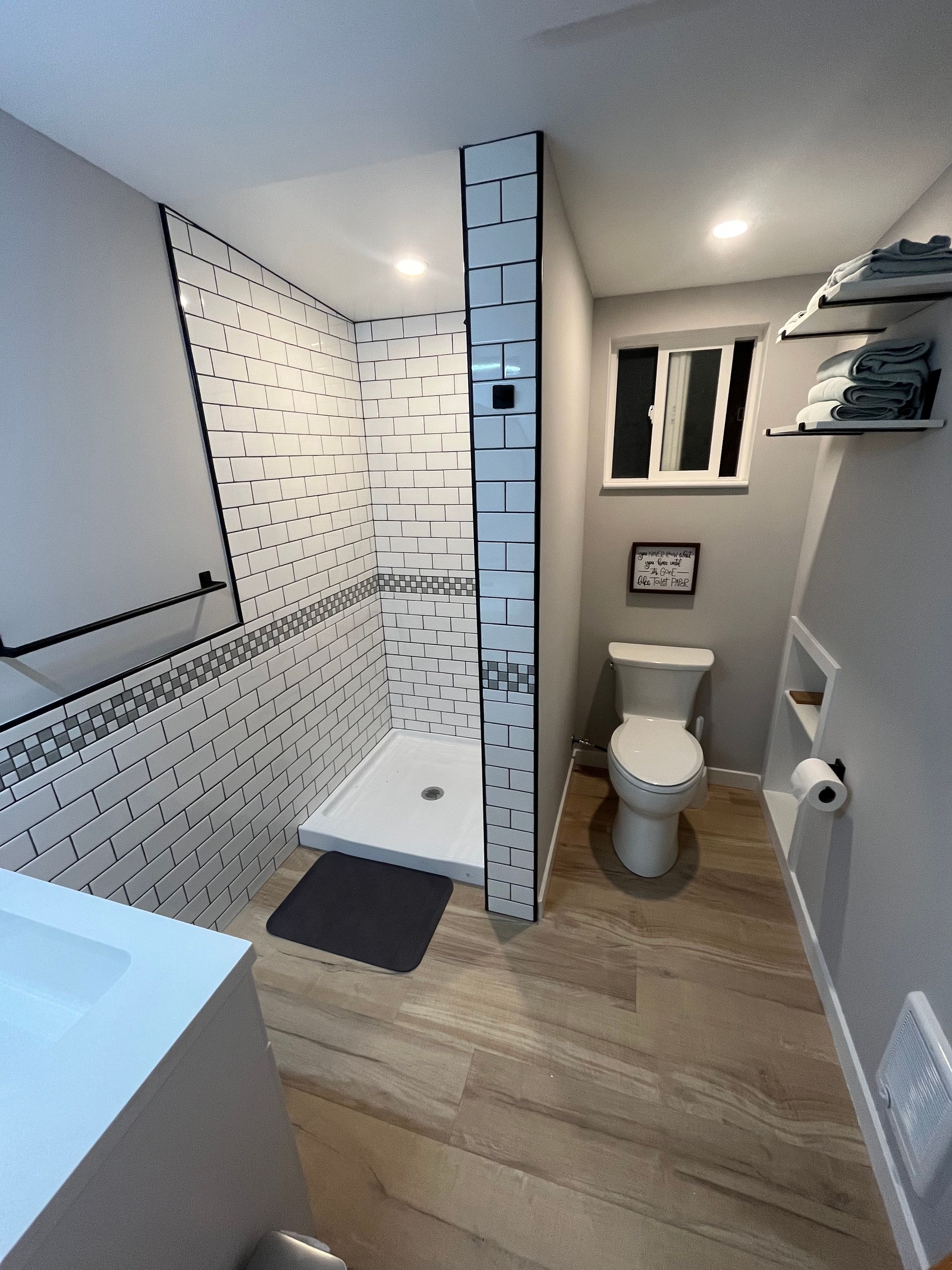 A modern bathroom with light wood floors, a tiled shower, a white toilet, and open shelving on a gray wall.