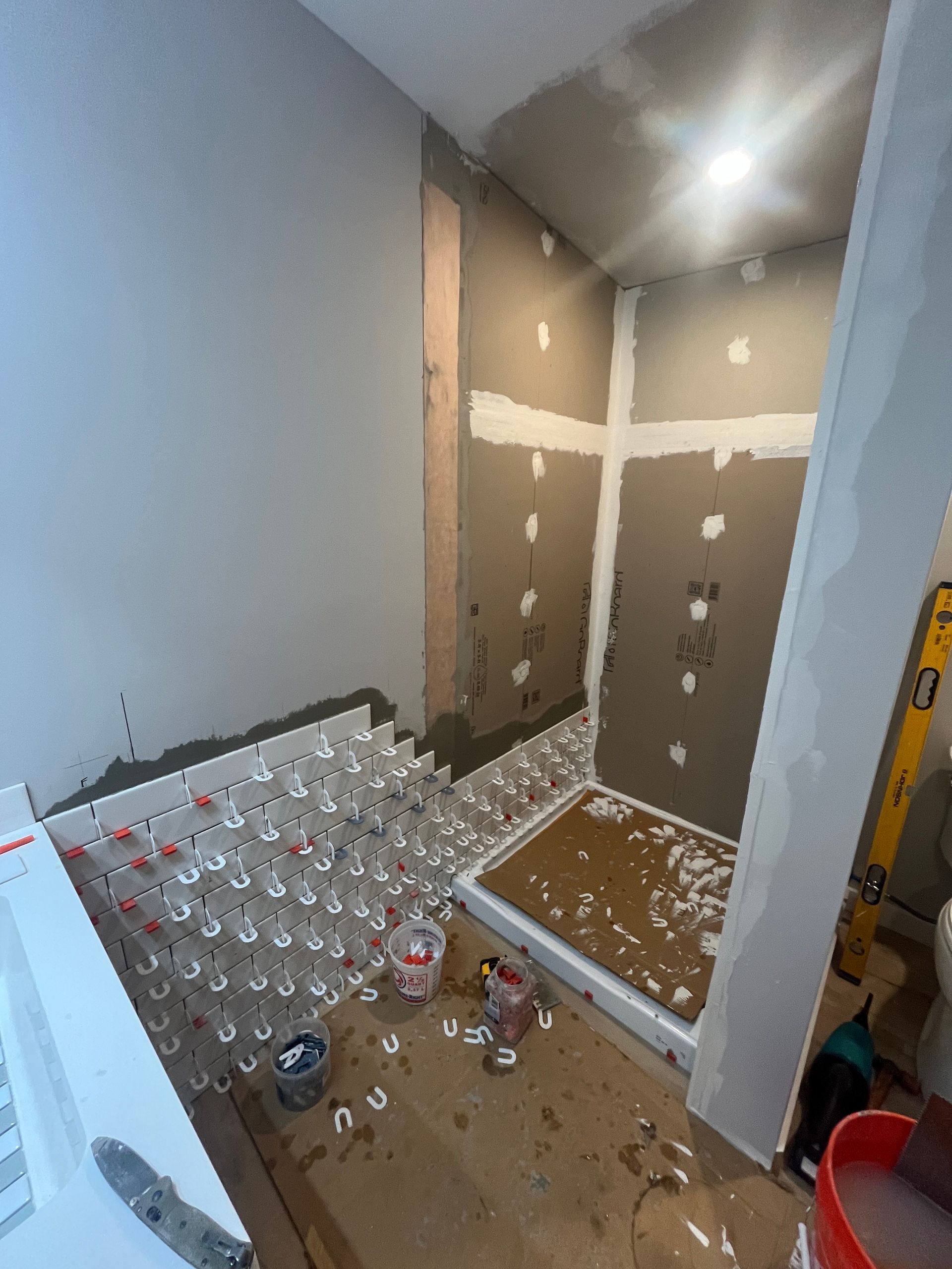 A shower enclosure under construction with white tiles installed along one wall using red leveling spacers.