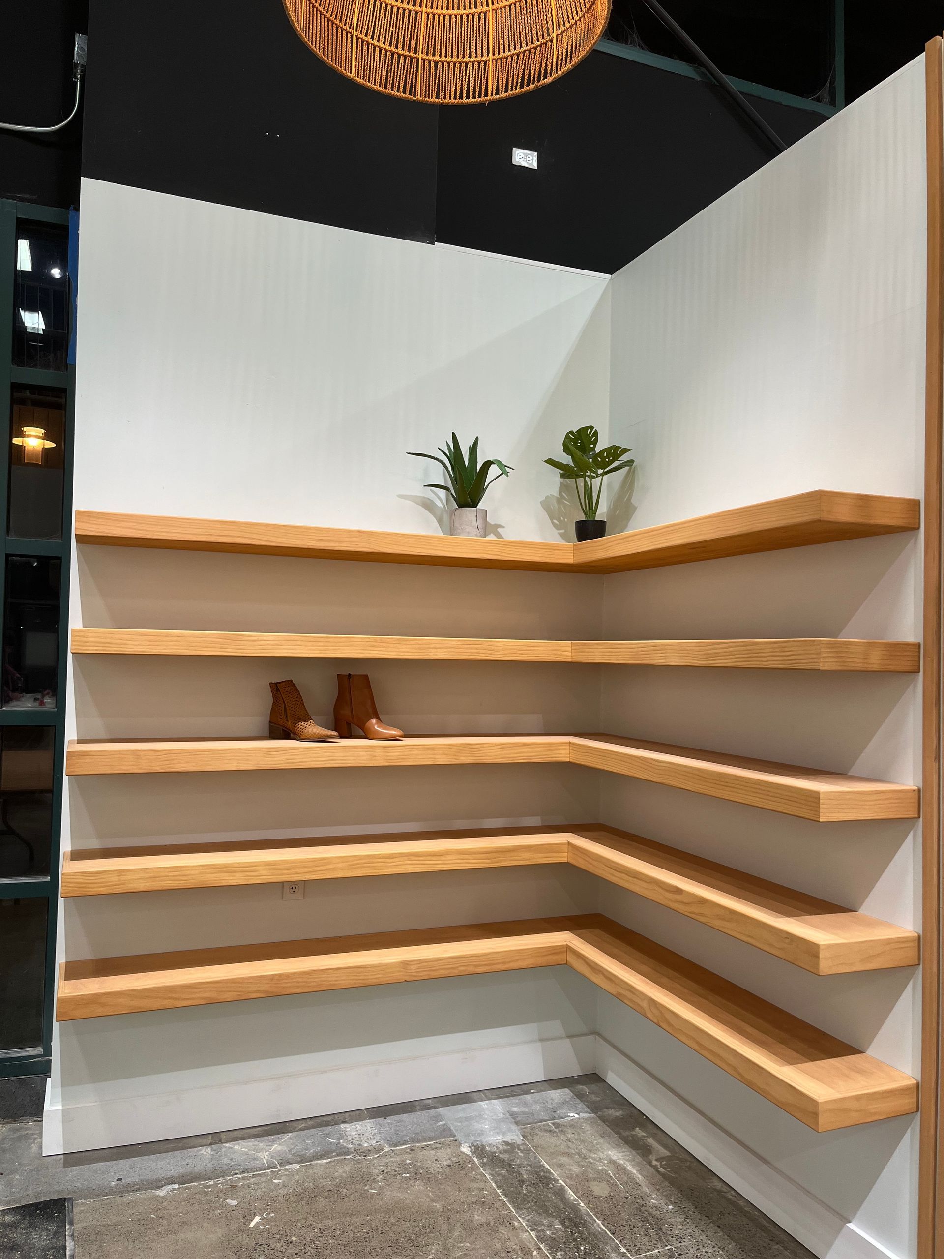A corner display features five stacked, light-wood floating shelves against white walls, with two small plants and boots.