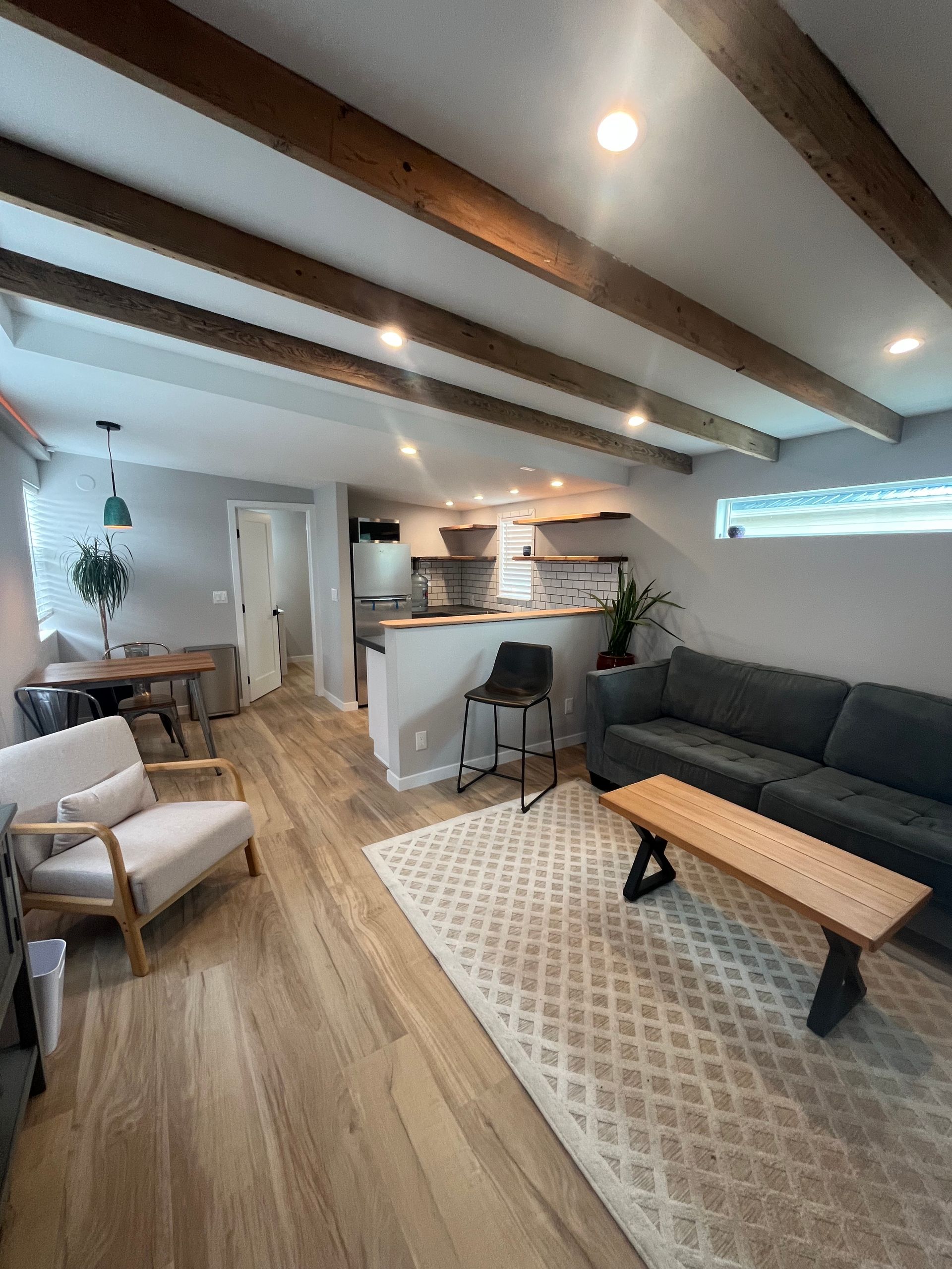 A modern living room with wood beams, a gray sofa, a light-colored accent chair, and a dining table, open to a kitchen.