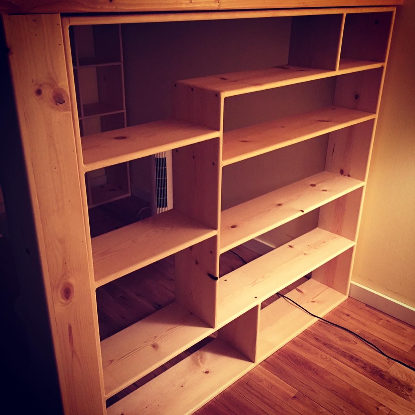 A large, unfinished wooden bookcase with staggered, asymmetrical shelves standing on a hardwood floor.