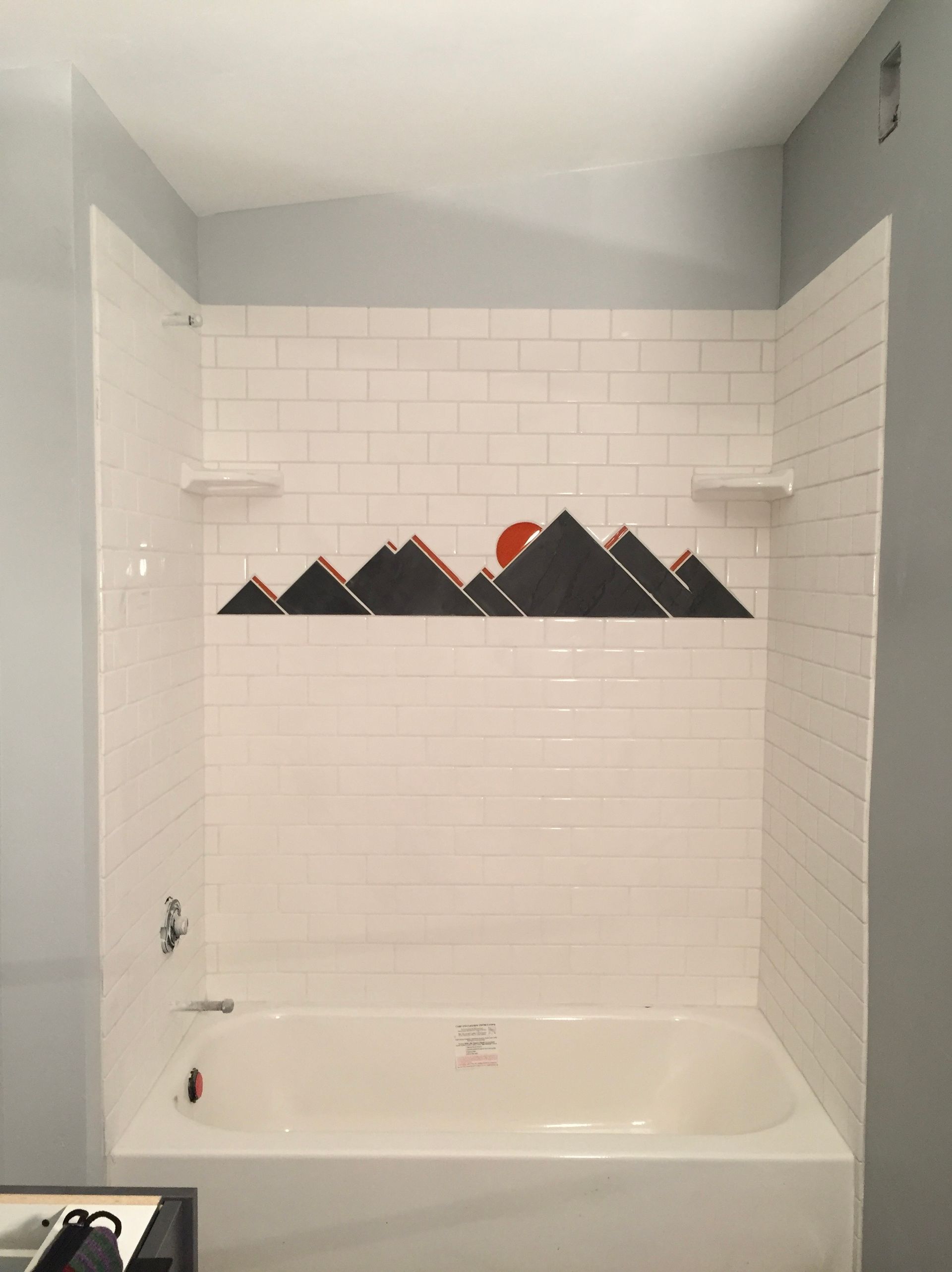 A white bathtub with subway-tiled walls featuring a decorative dark gray mountain range mural and a red sun.
