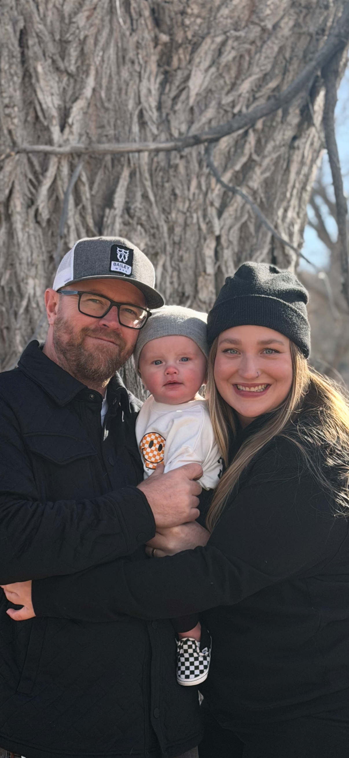 A family of three smiling outdoors in front of a large tree. They are wearing hats and casual dark clothing.