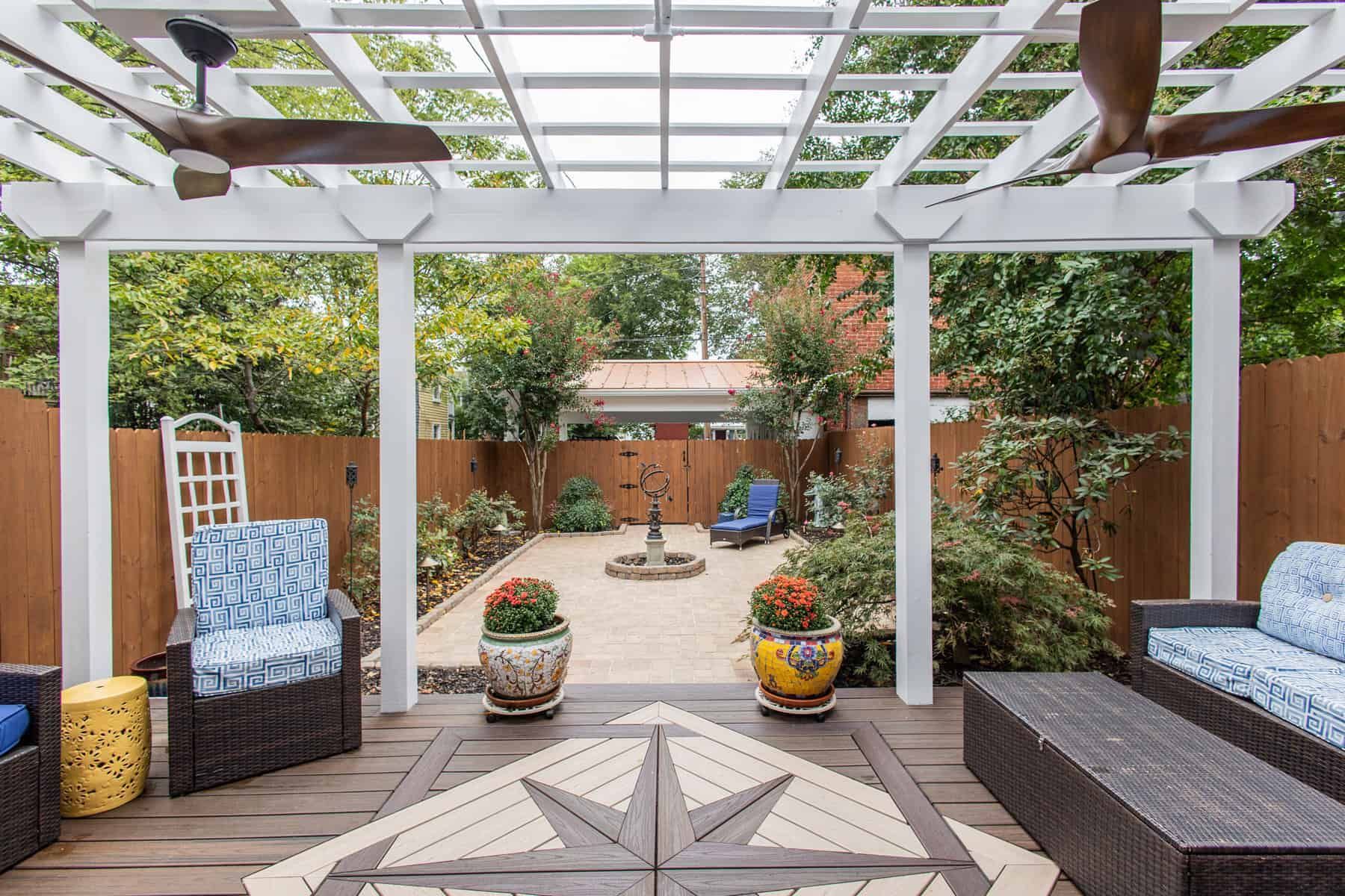 Outdoor patio with pergola, seating, and decorative tile. Garden view with fountain, trees, and wooden fence.