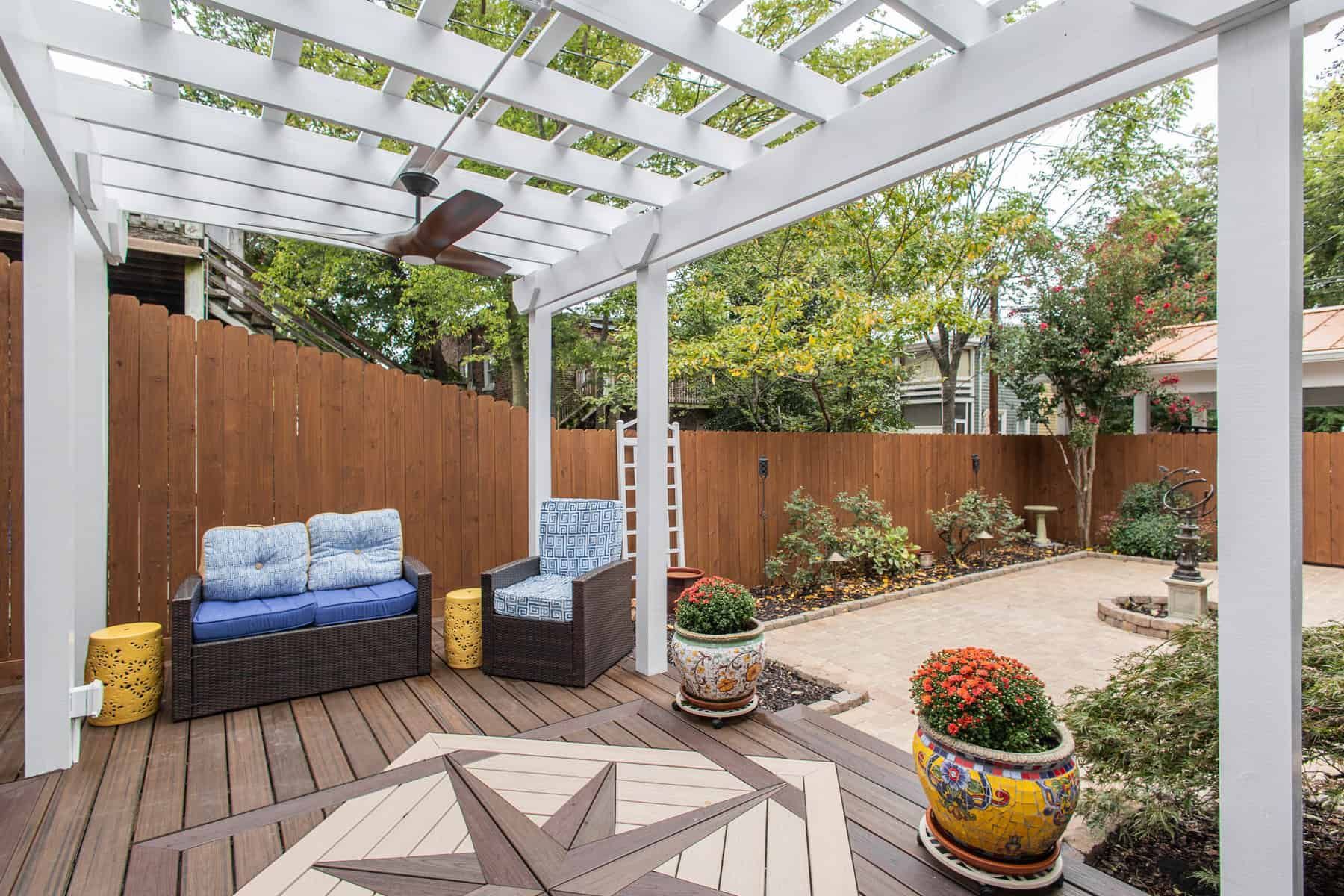 Patio with pergola, furniture, wood deck, brown fence, plants, and a ceiling fan.