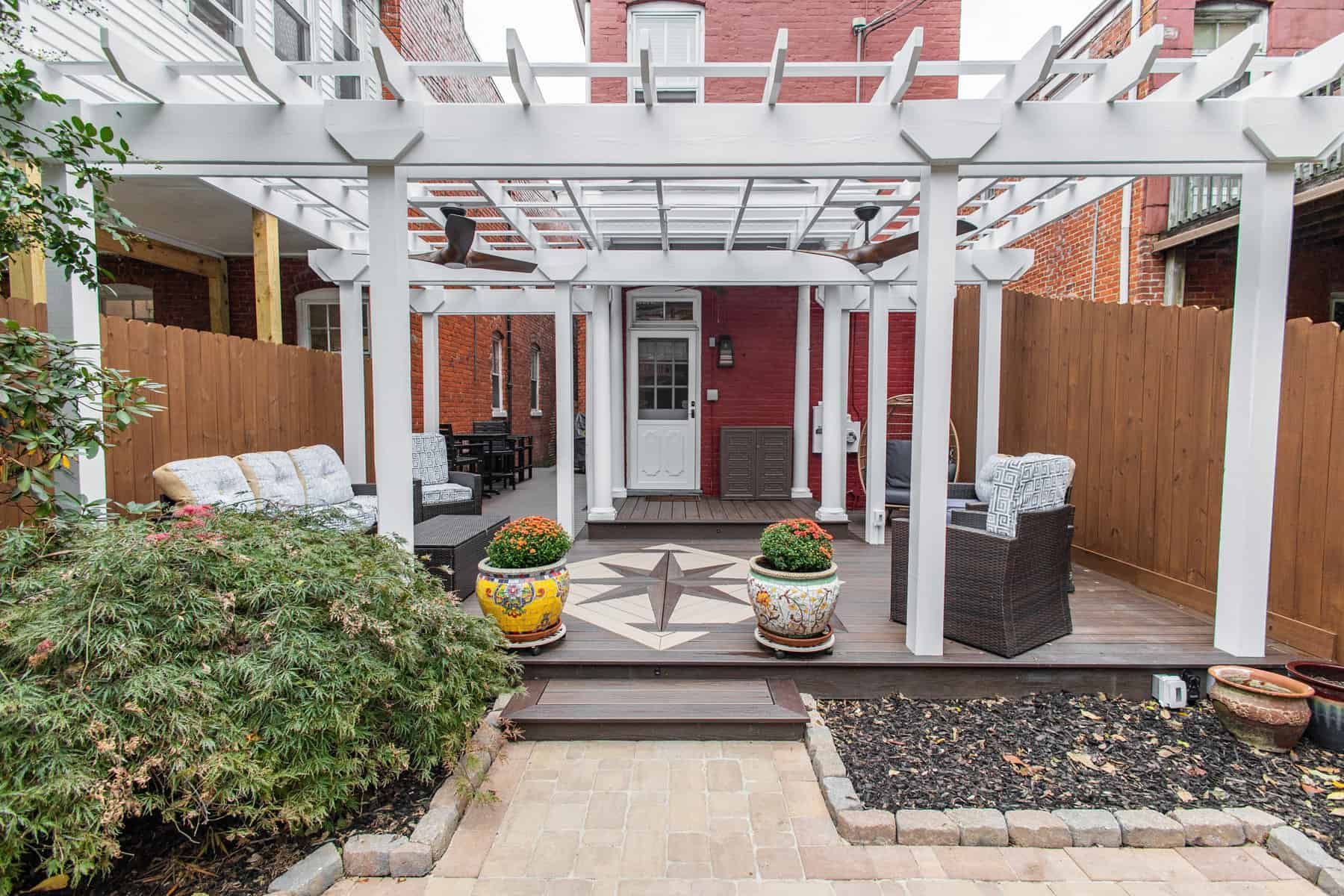White pergola over a backyard patio with seating, potted plants, and a door to a red brick building.
