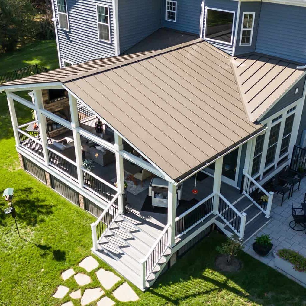 A two-story house with a screened-in porch and tan metal roof, white railing and trim, and steps leading to a paved patio.