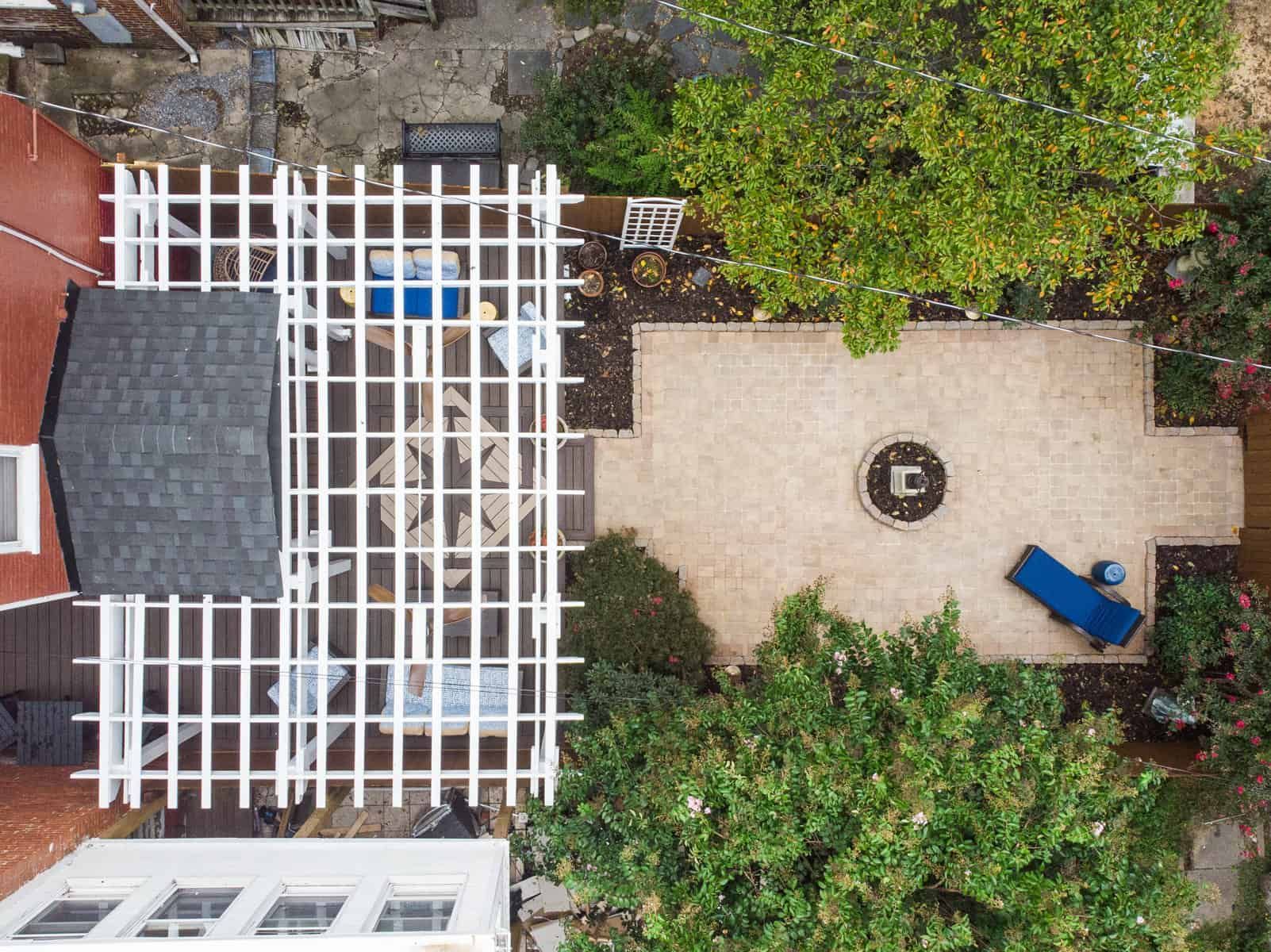Overhead view of a backyard patio with a white lattice pergola, a stone fire pit, and surrounding greenery.