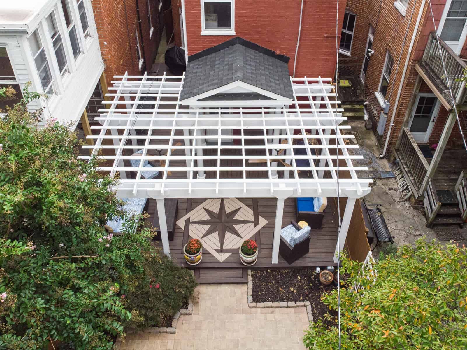 Overhead view of a backyard patio with a white pergola, dark wood deck, and a small, gabled roof structure.