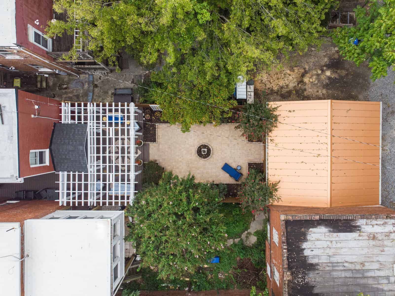 Overhead view of a backyard patio with trees, a fire pit, and a pergola. Buildings surround it.