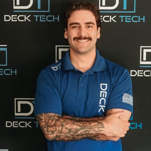 Man with mustache in blue polo shirt with arms crossed, standing in front of a backdrop with