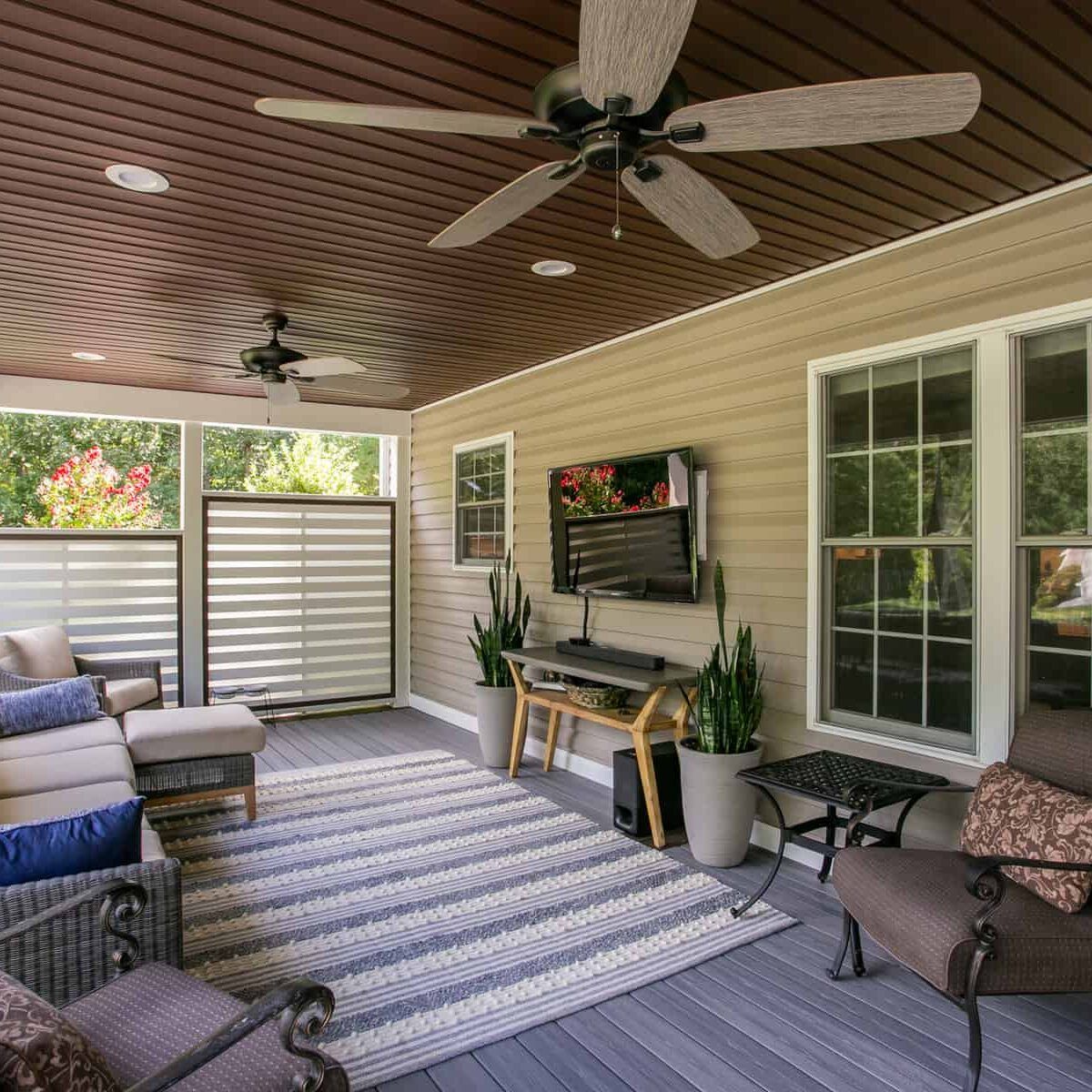 Enclosed outdoor living space with couch, rug, TV, plants, and ceiling fans.