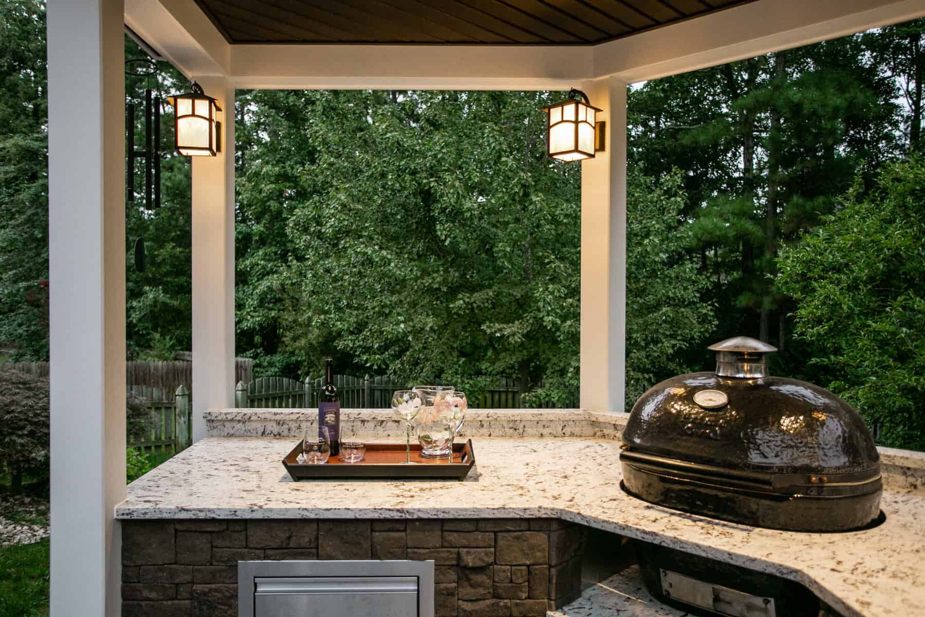 Outdoor kitchen with grill, counter, and overhead lighting. Lush green trees in background.