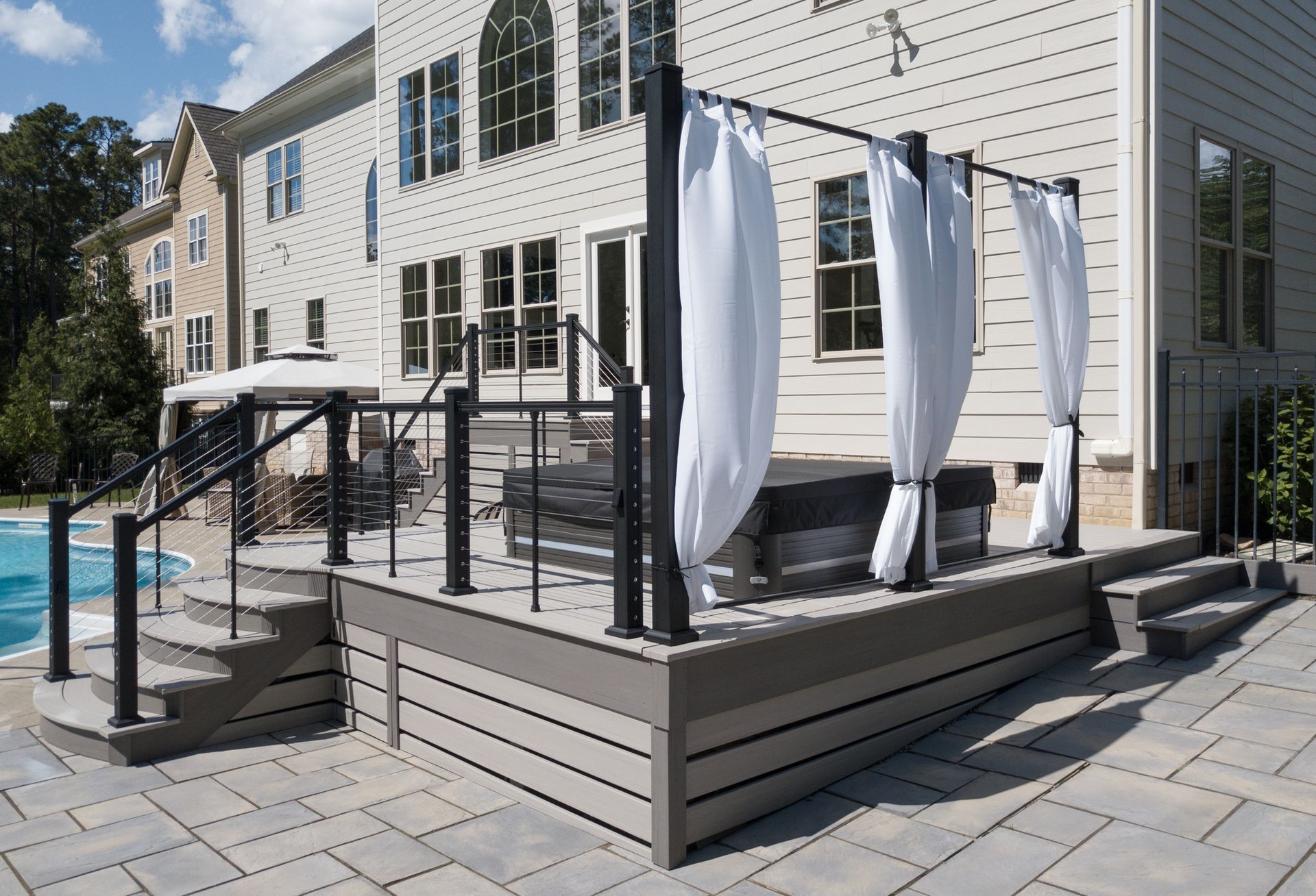 Patio deck with hot tub, curtains, steps, and pool in front of a house.