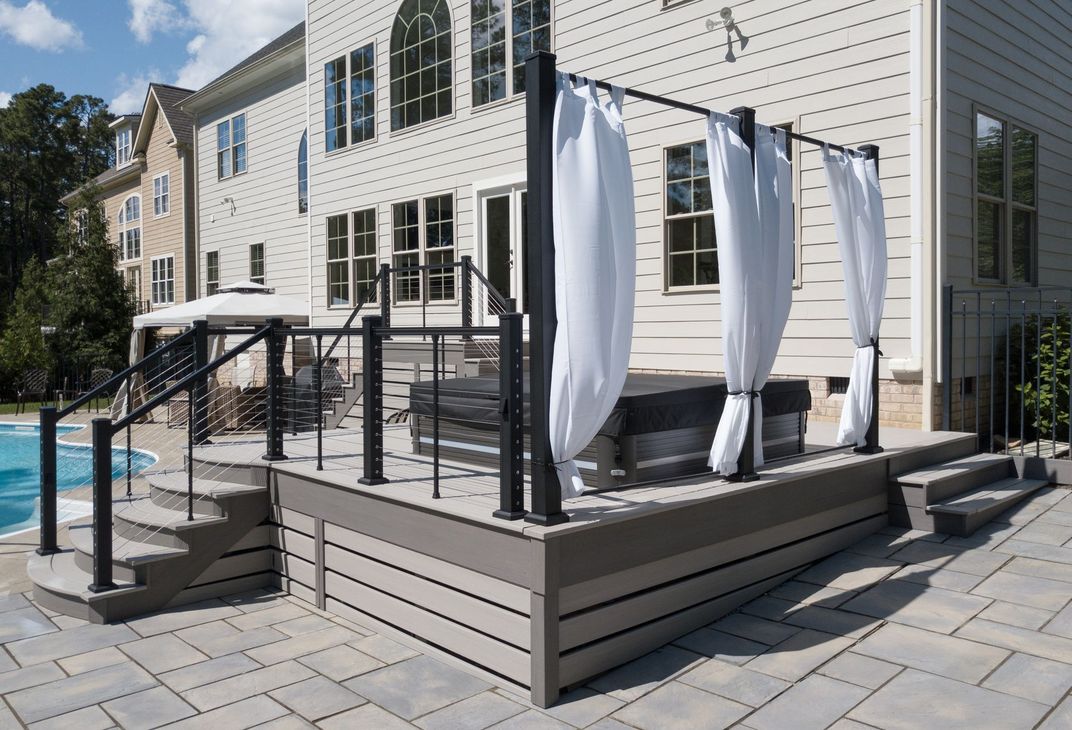Outdoor deck with hot tub, steps, and draped curtains, next to a pool and house.