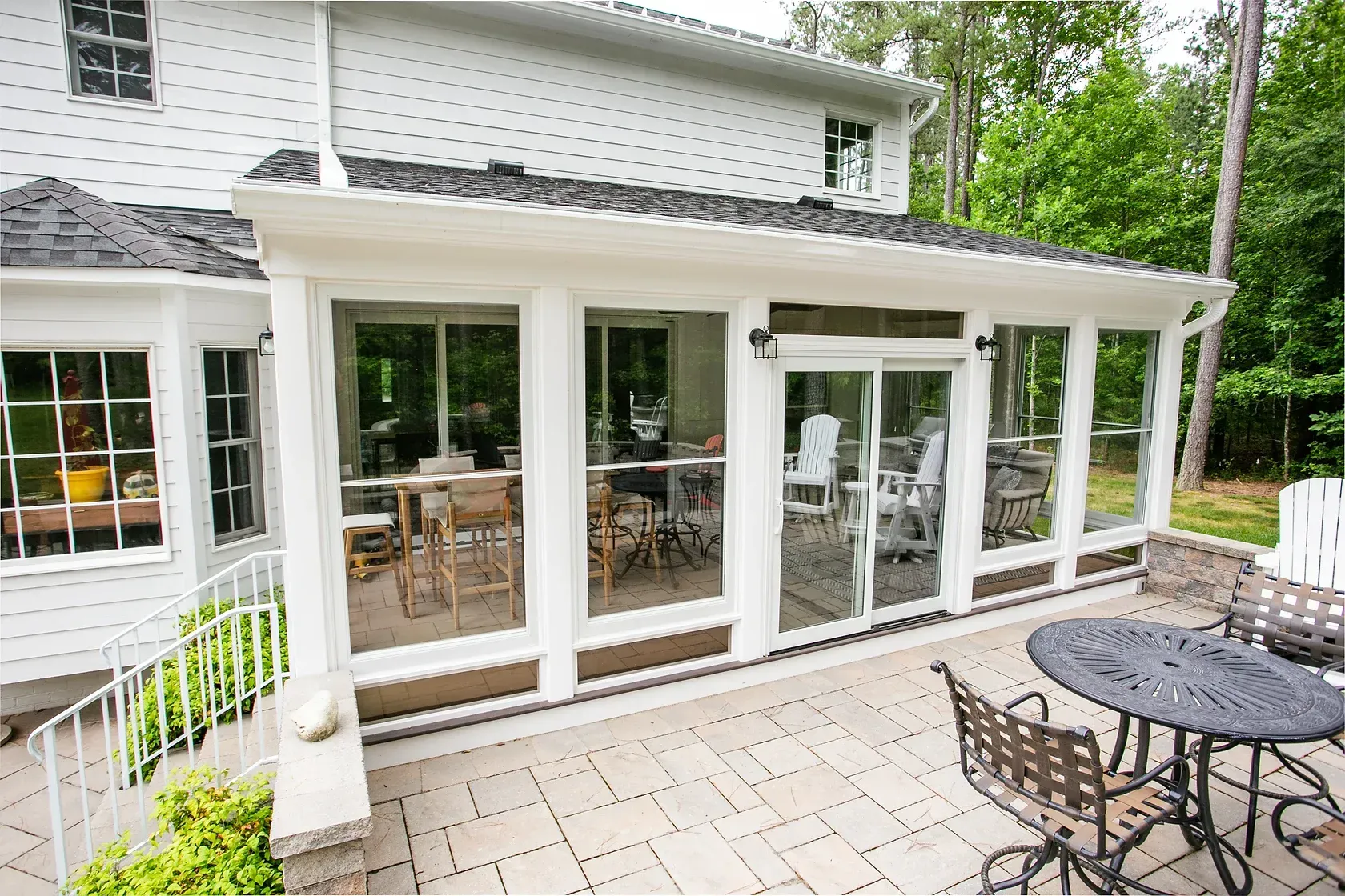 Exterior of a home with a patio and sunroom with large windows, surrounded by trees.