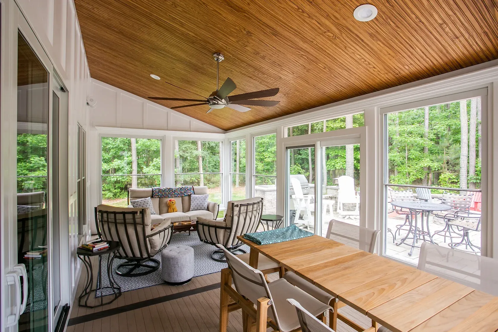 Sunroom with wood ceiling, large windows, and outdoor furniture overlooking a patio and trees.